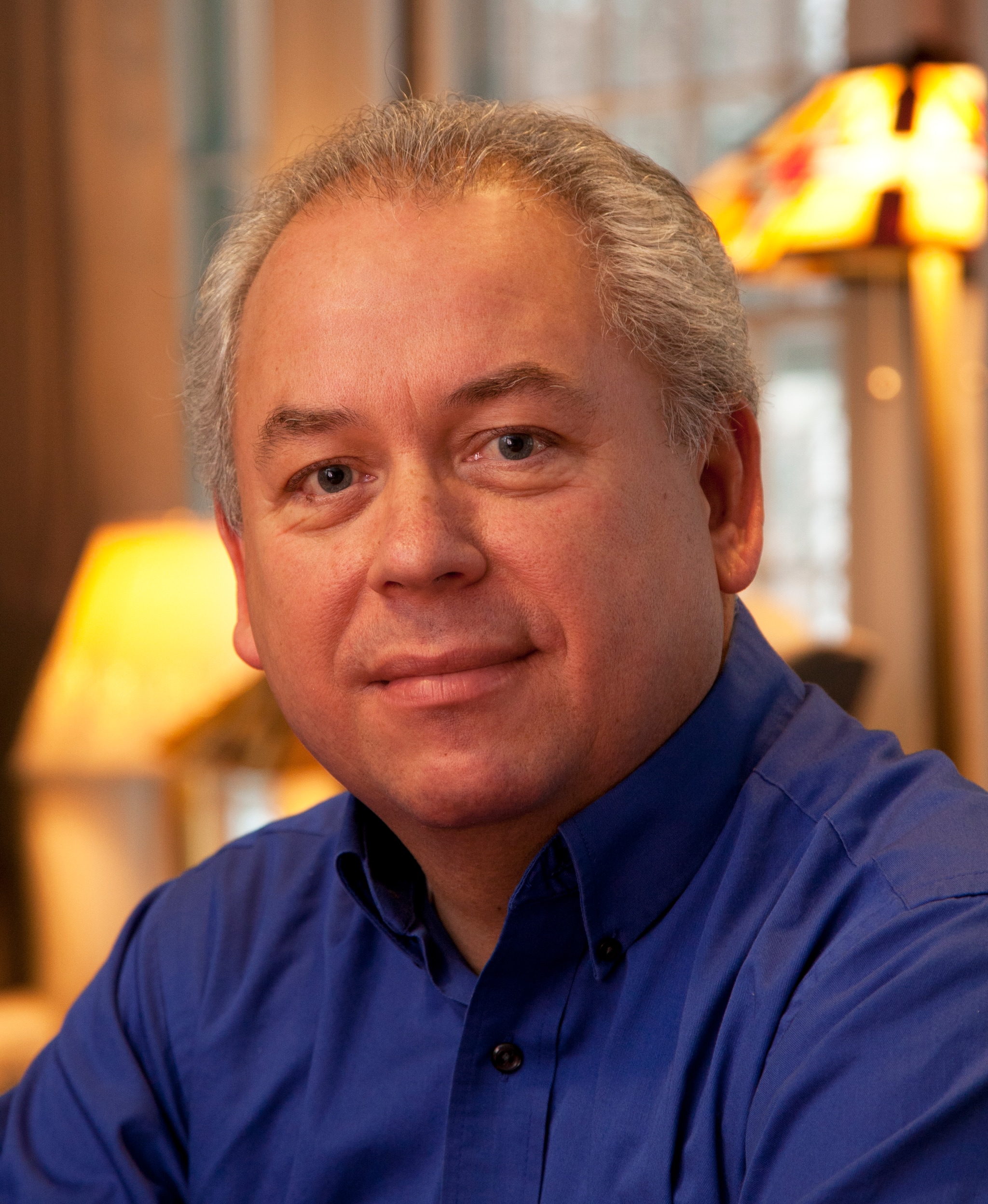 A man in a royal blue button down shirt sitting with lamps glowing behind him. He has carefully combed graying hair and smiles slightly.