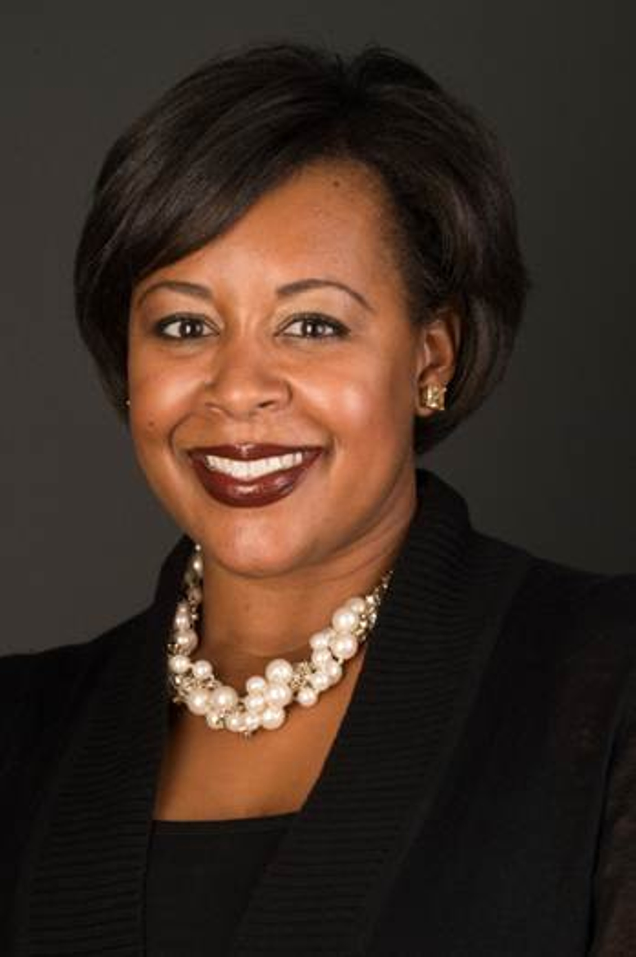 A photo of Kriston Alford McIntosh posing against a dark gray background. McIntosh smiles and has hair parted on the left side of her head with bangs in a diagonal across her forehead. She wears a heavy white beaded necklace and high collared black jacket. 