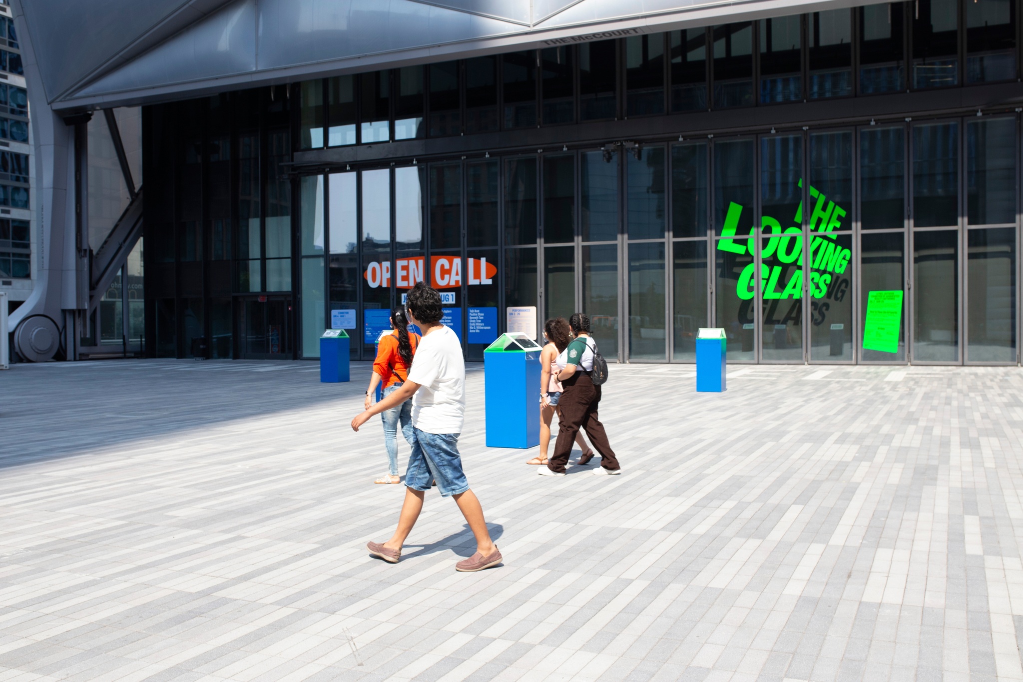 A group of four people walking across a sunlit plaza at a distance from each other with bright blue plinths dotting the space around them