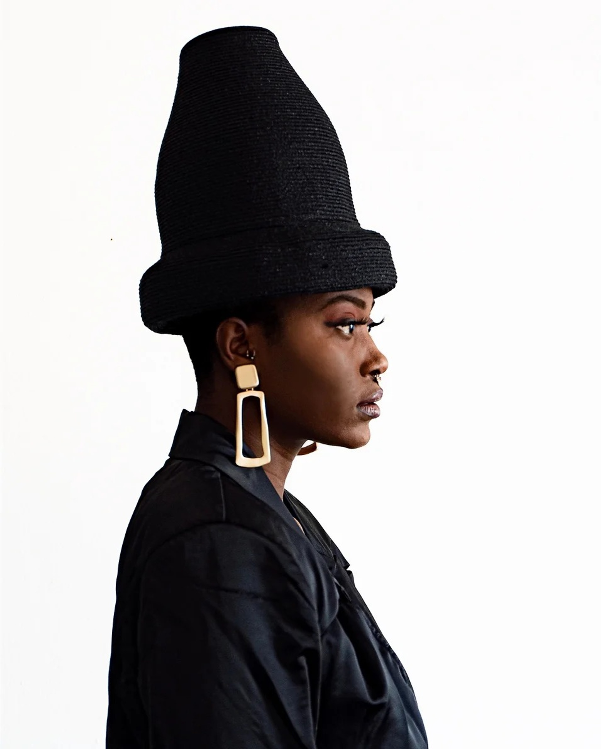 A portrait of artist Chelsea Odufu, seen in profile against a stark white background. Chelsea is a Black woman who wears a dark jacket, large rectangular gold earrings, and a tall, dark conical hat with a narrow boxy brim. 