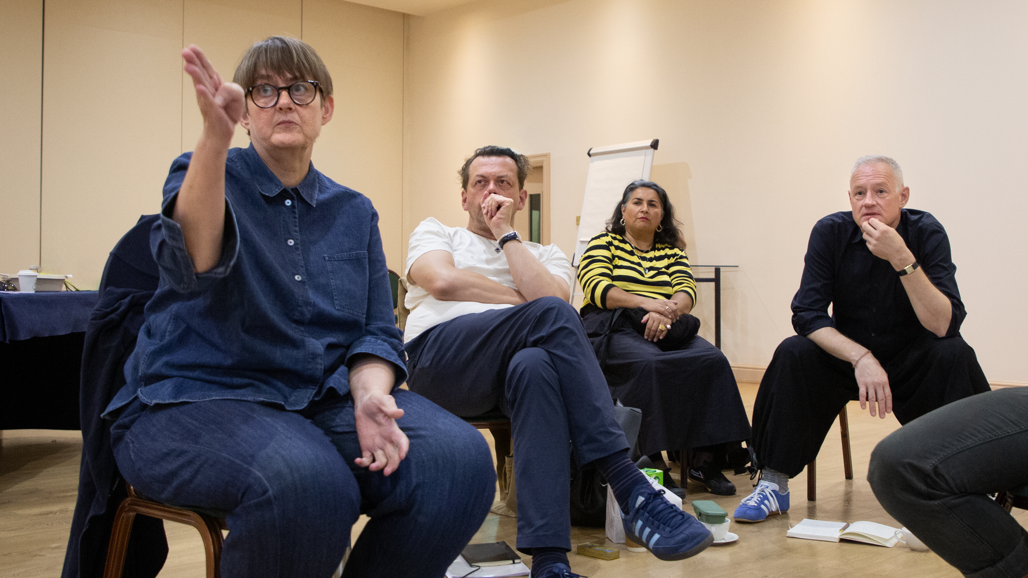 The director Sarah Frankcom, writer Simon Stephens, casting director Shaheen Baig, and producer Todd Eckert sit in a row in a rehearsal room. They all train their attention toward us. Sarah is speaking with one hand gesturing in front of her. 