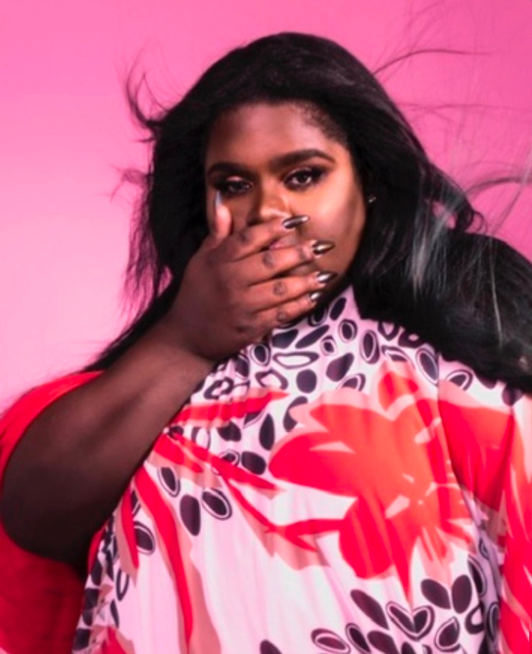 A portrait of Precious against a pink backdrop. Precious wears a floral print dress and holds one hand up to their face.