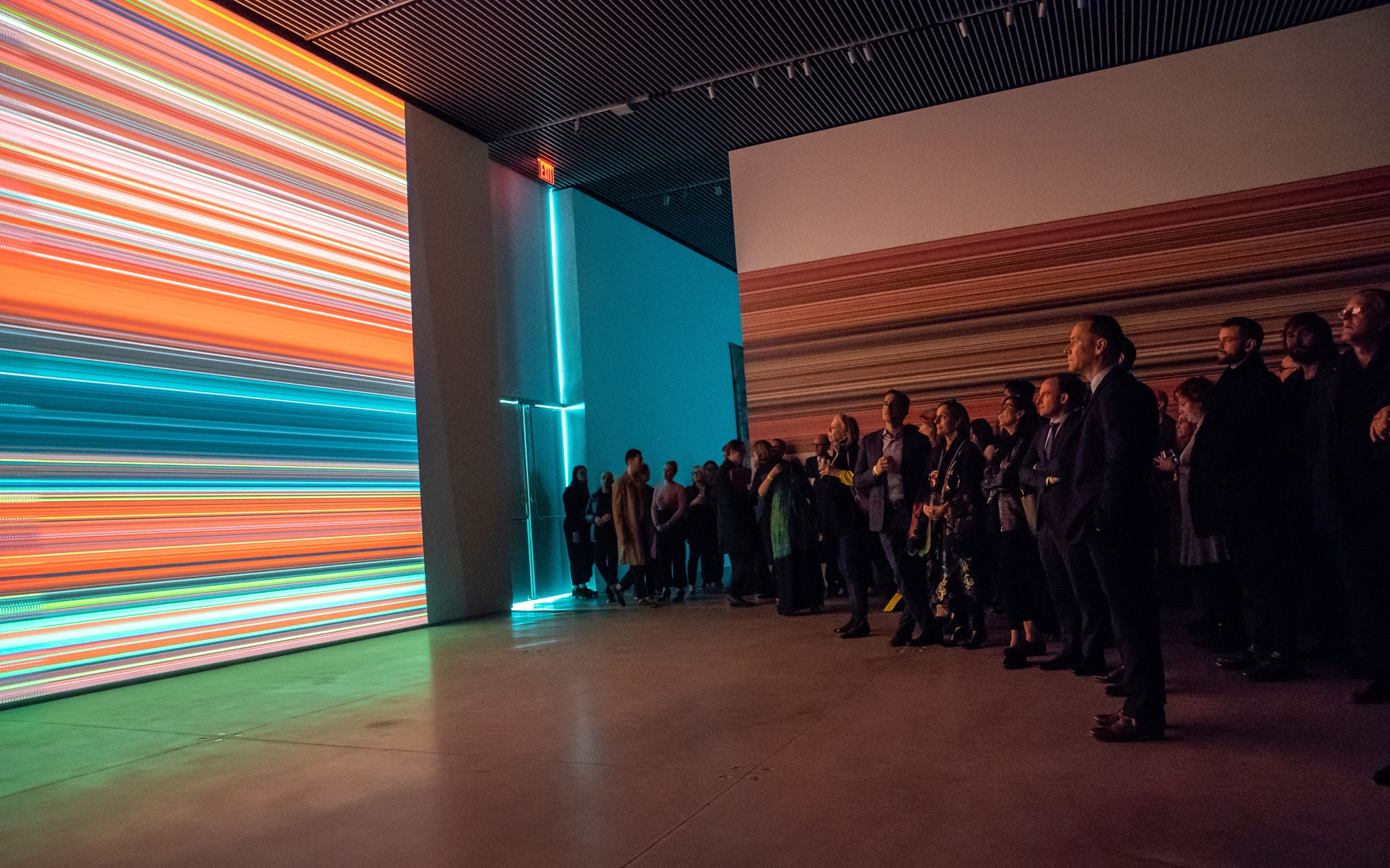An audience in a gallery is gathered in front of a film projected on a white wall. The film displays a colorful abstract pattern of stripes. 