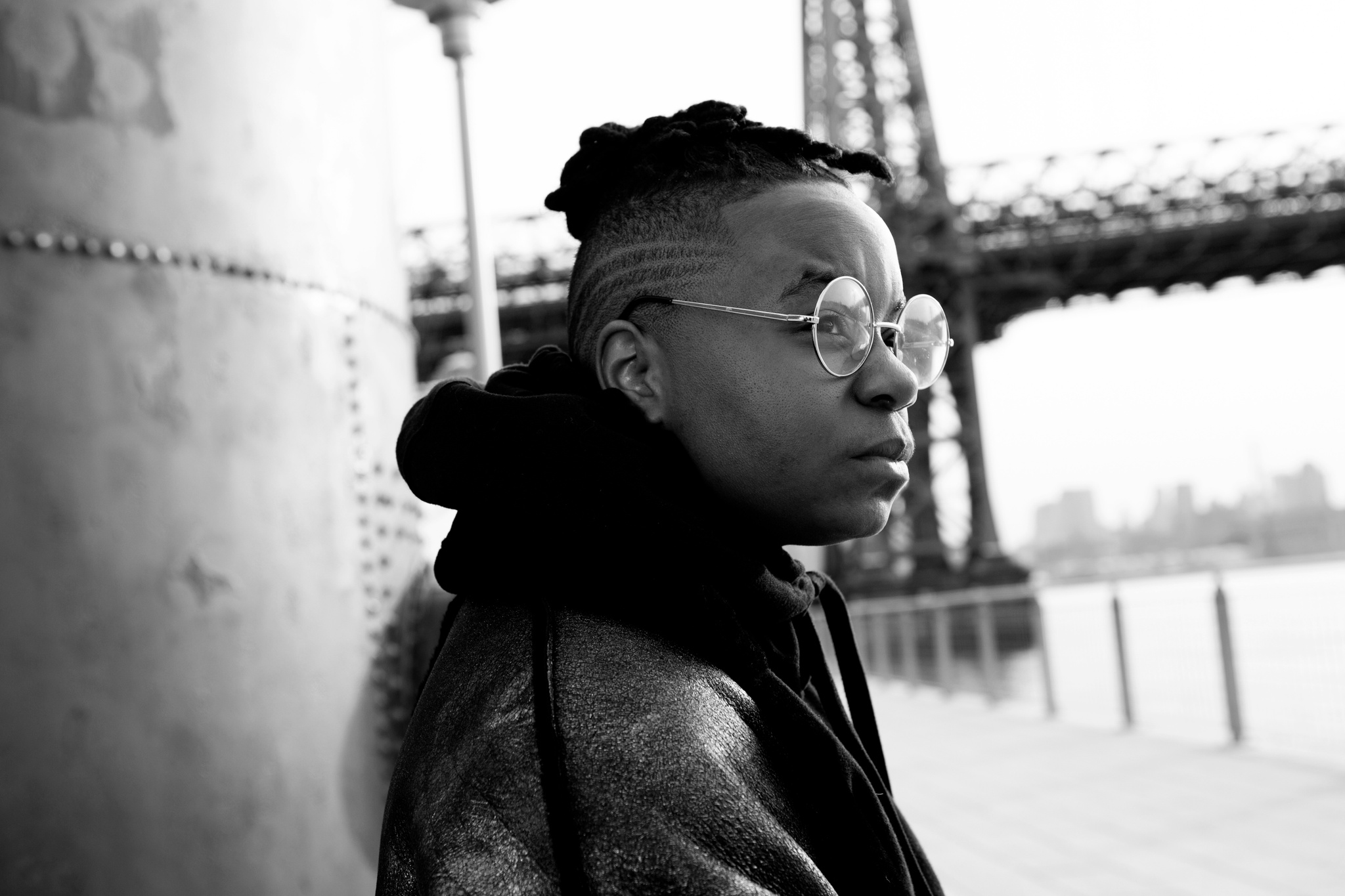 A black-and-white photo of Kei Williams. They are leaning against a metal support looking out at a river with a tall bridge crossing the image in the background. 