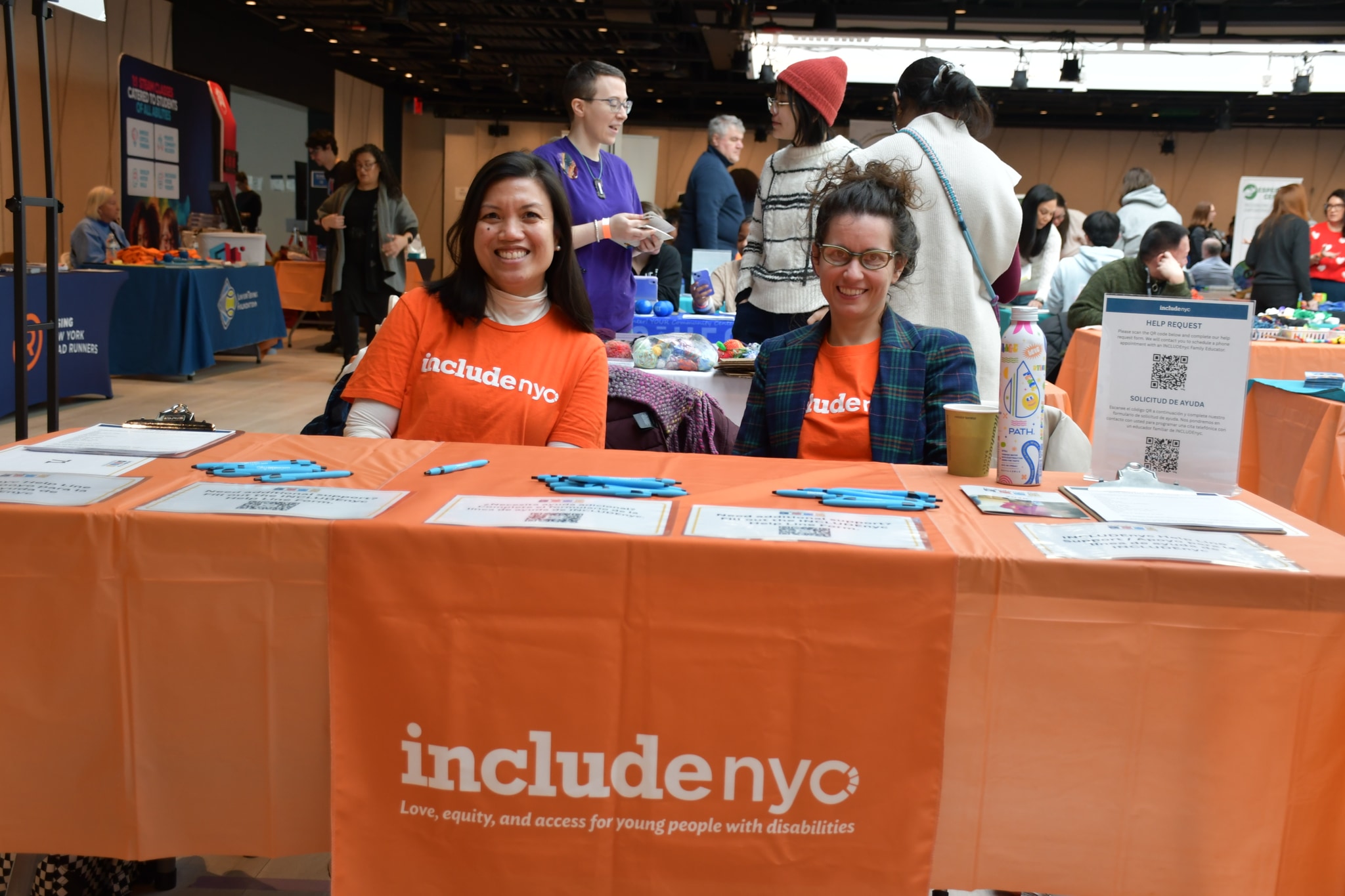 Two women, one Asian and one white, sit behind a table draped in a bright orange cloth that reads "Include NYC: Love, equity, and access for young people with disabilities." They both smile directly at us.