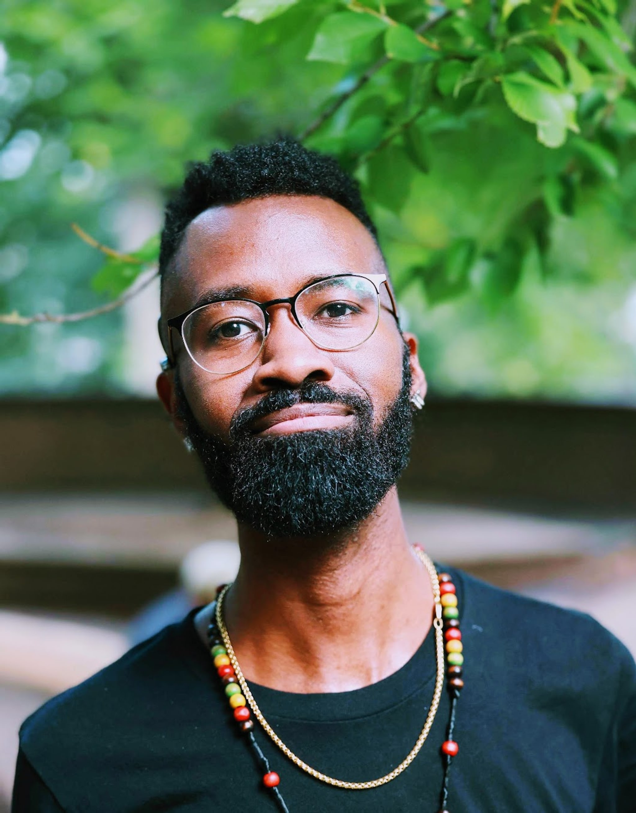 A portrait of Wade Green, a Black Deaf actor, who poses outside beneath a leafy green branch of a tree. He is seen from the chest up wearing a black t-shirt and glasses. He has a full beard and short hair. He looks directly at us with a satisfied look on his face. 