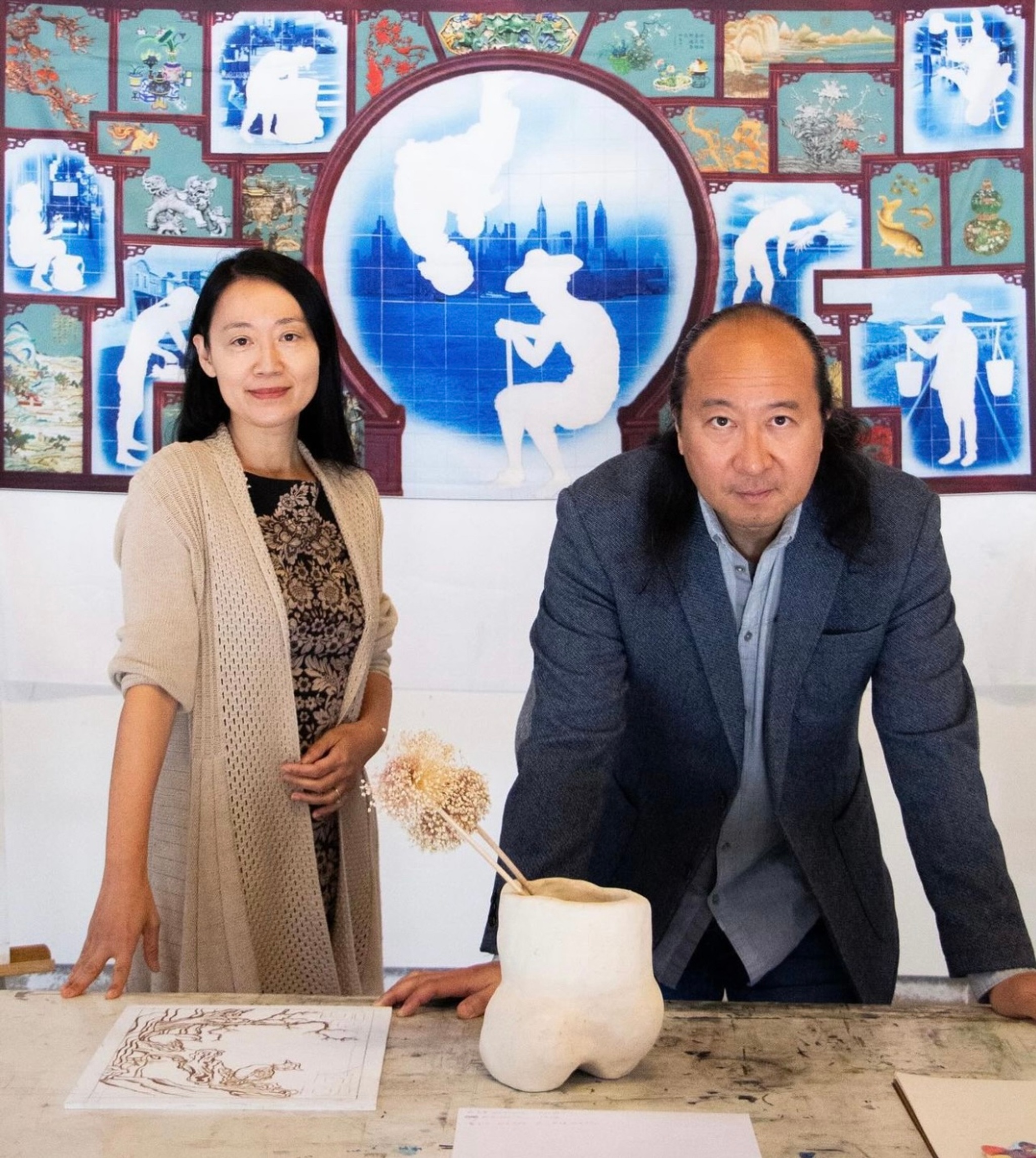 A portrait of artist duo Lily Honglei. Lily Yang, a Chinese woman, stands to the left of the photo in a long beige cardigan. She looks directly at us smiling and reaches down with one hand to a work table in front of her. Beside her, Chinese artist Honglei Li leans on the same table with both arms. He wears a blue blazer over a light blue shirt.