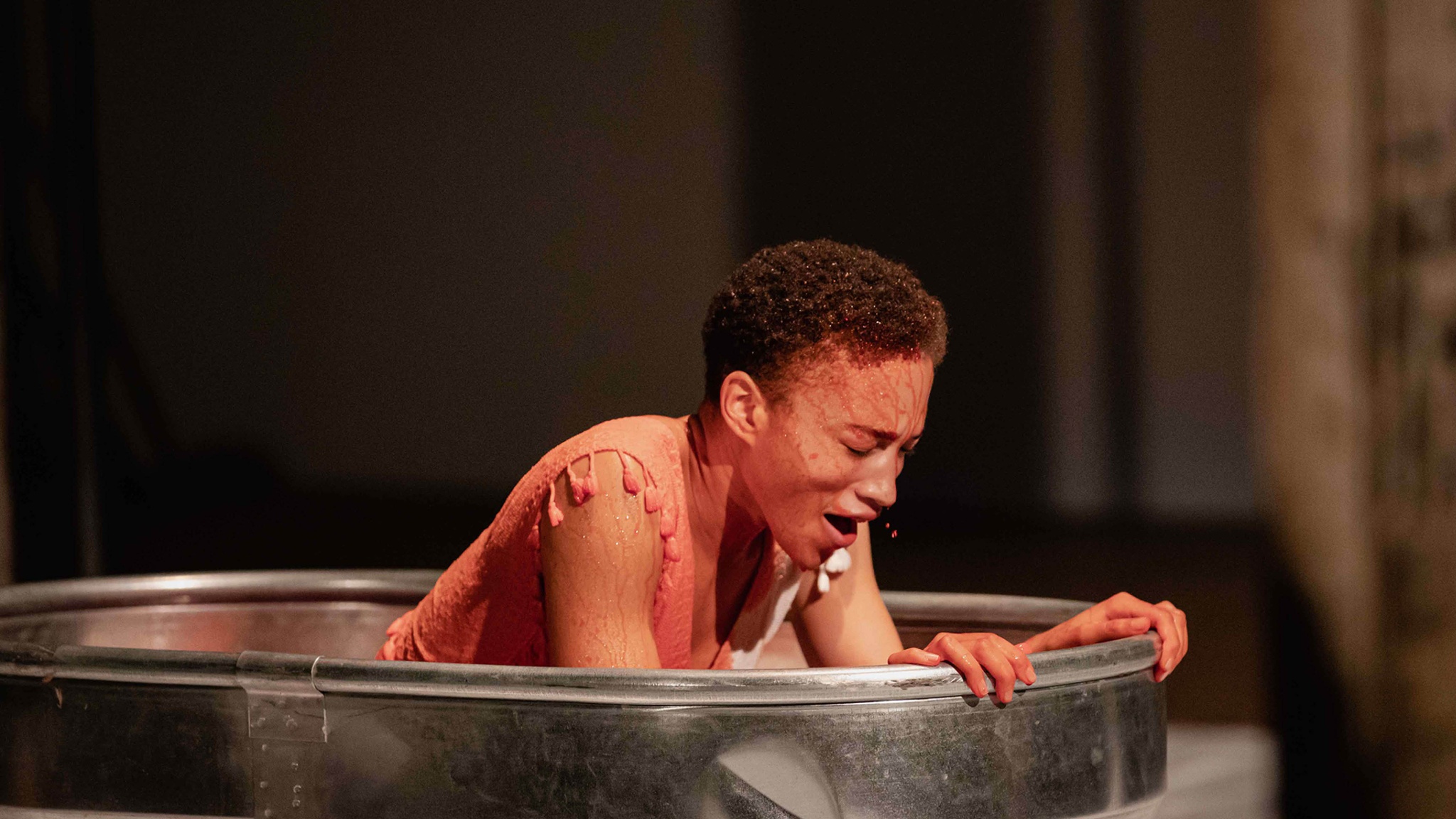 The artist Asia Stewart rises from the red water inside a metal wash tub, gasping for breath with eyes closed. The red liquid pearls in her hair and runs in rivulets down her face, dying the sleeveless white dress she wears red.