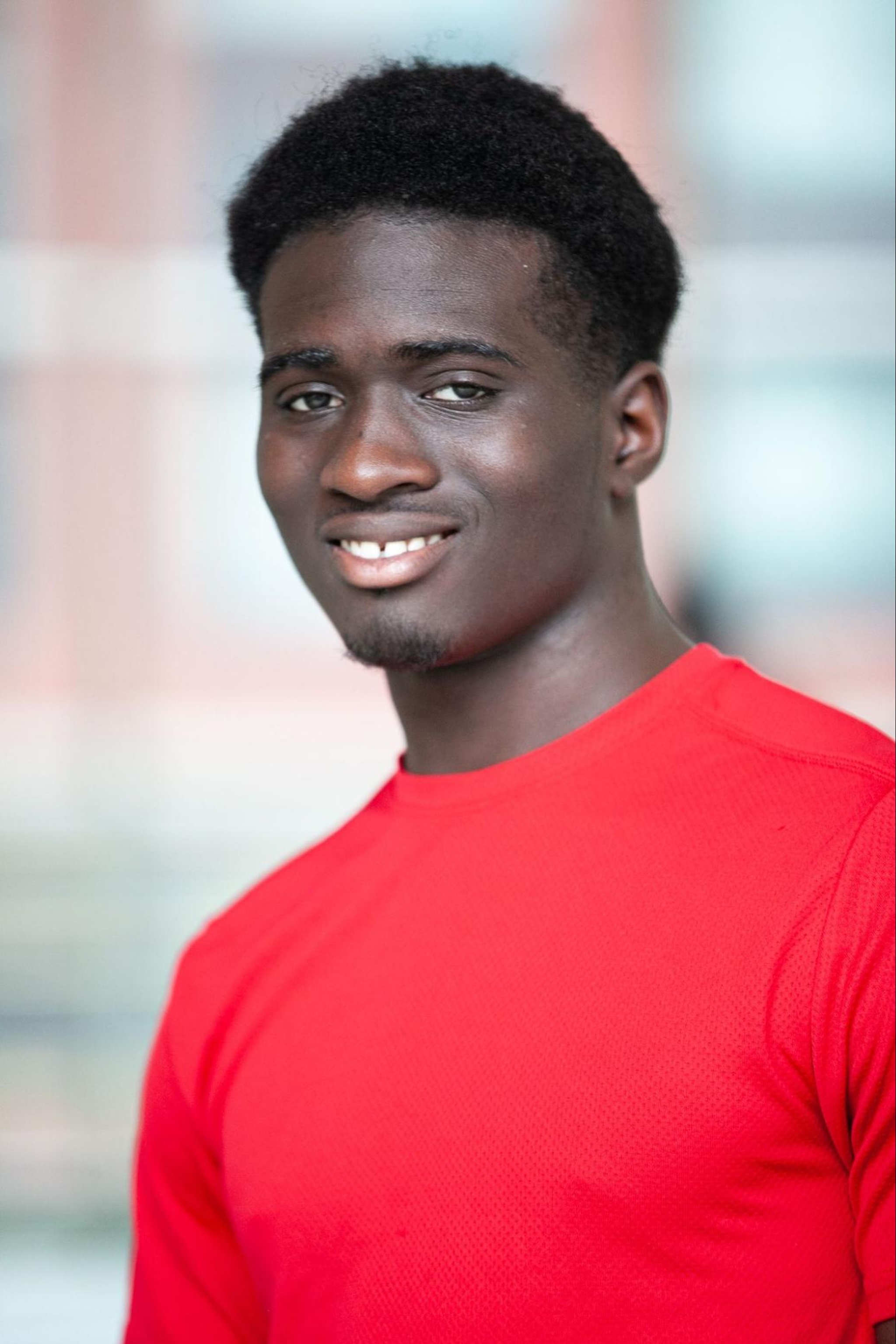 A portrait of dancer Malachi Kingston, a Black man with dark brown skin who wears a bright red t-shirt. He looks directly at us, smiling. 