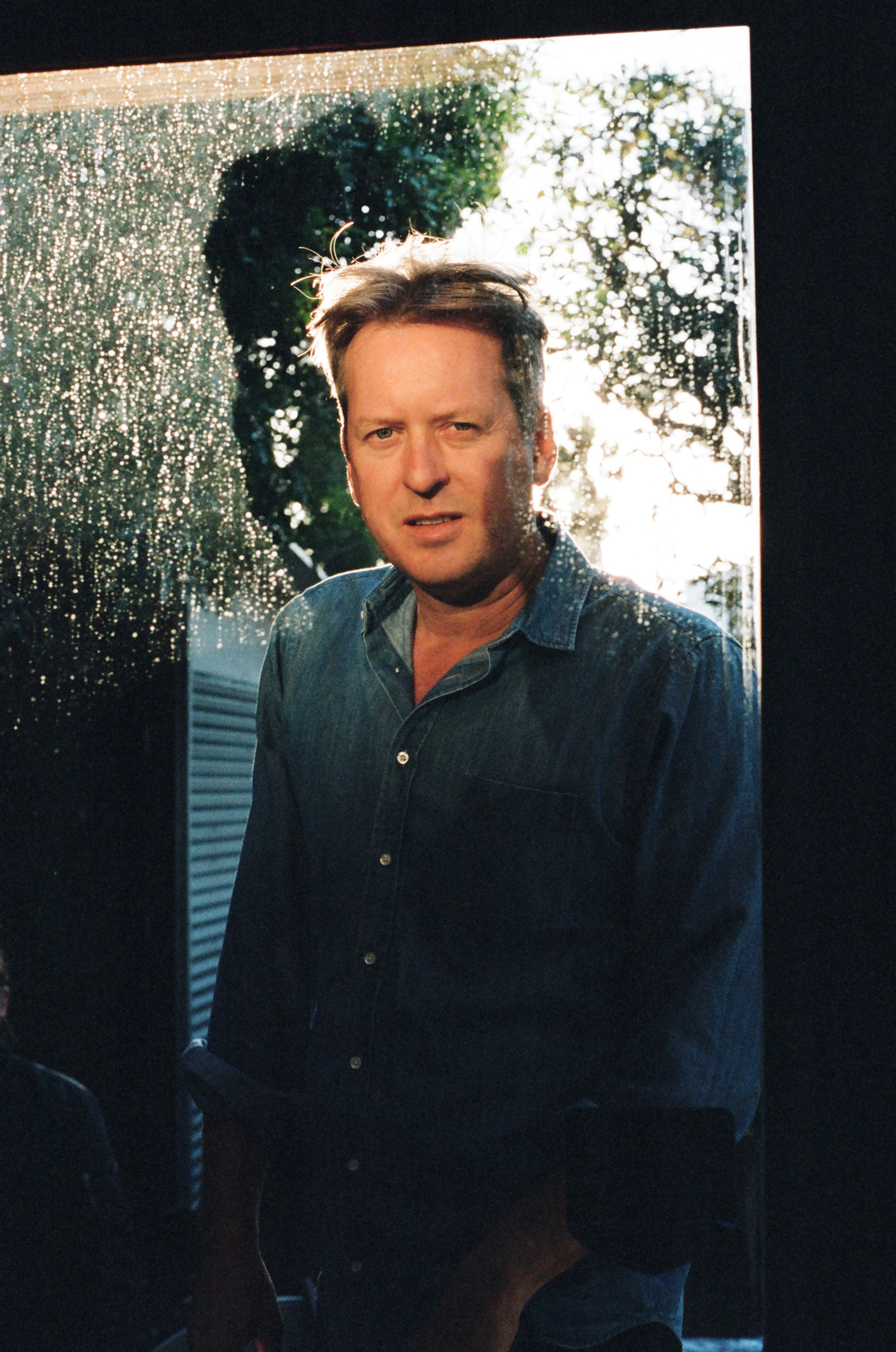 A portrait of artist Doug Aitken, a white man with short brown hair parted to the side. Doug peers at us through a clear windowpane, speckled with drops of rain. Bright sunshine illuminates him from behind. 