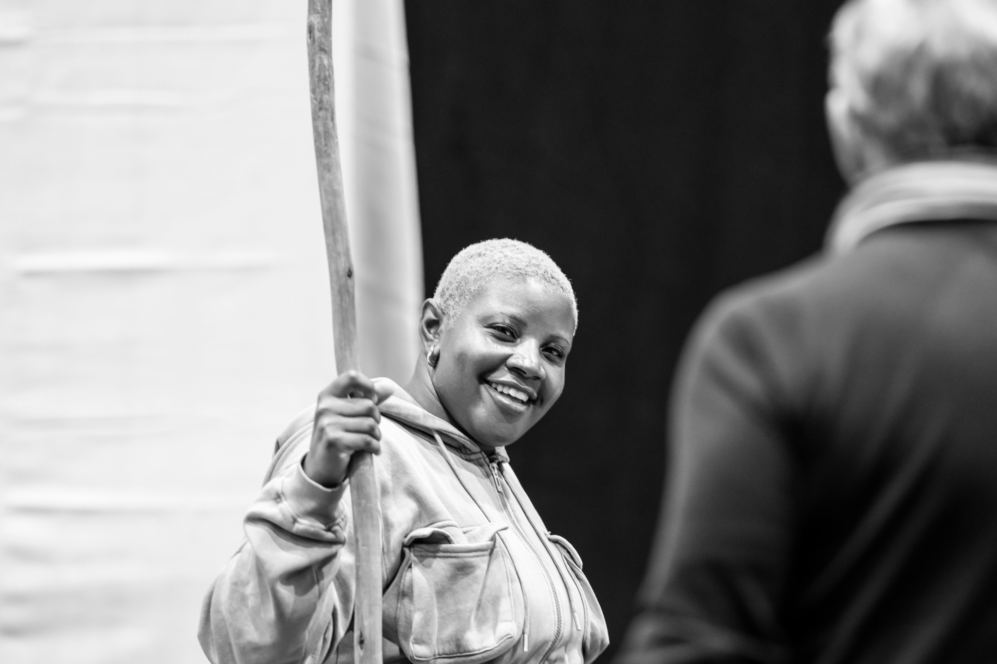 Actor Mara Allen, a Black woman with a short shaved hair dyed blond, holds a prop staff and smiles broadly during rehearsal. She is looking at another member of the cast whose back is turned toward us.