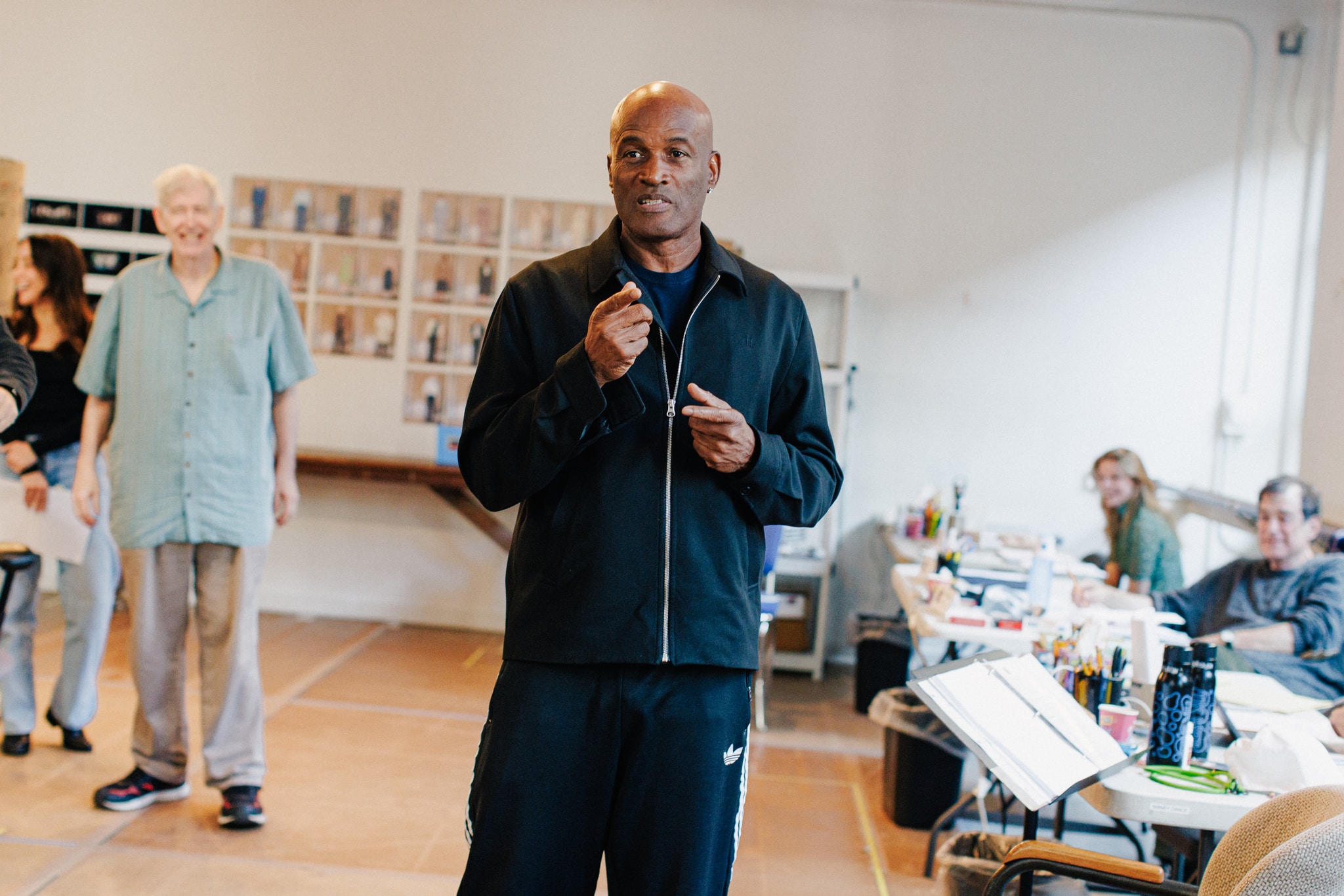 A black and white photo of director Kenny Leon. Kenny is a middle aged black man who is in mid-sentence addressing the rehearsal space in which he stands. His hands are raised in front of him as he speaks.