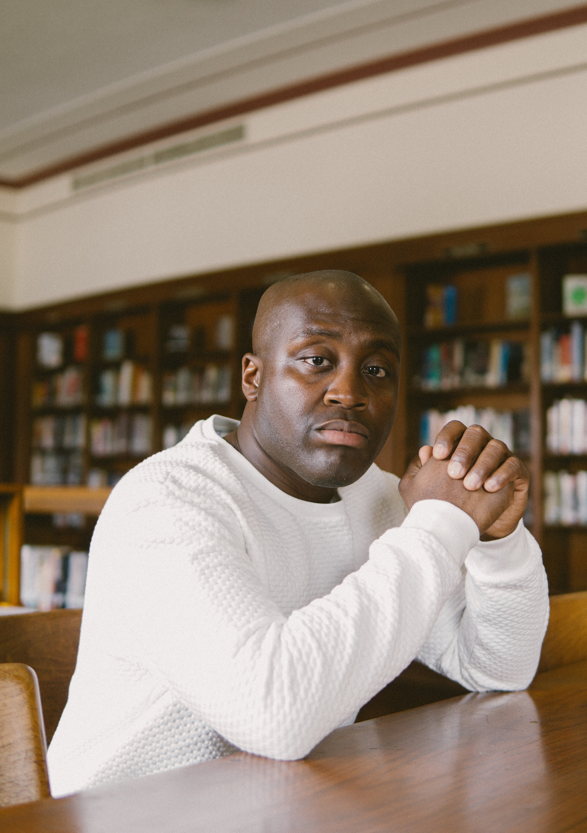 A photo of Larry Ossei-Mensah sitting at a library table with elbows on the table and hands held up and clasped in front of him. 