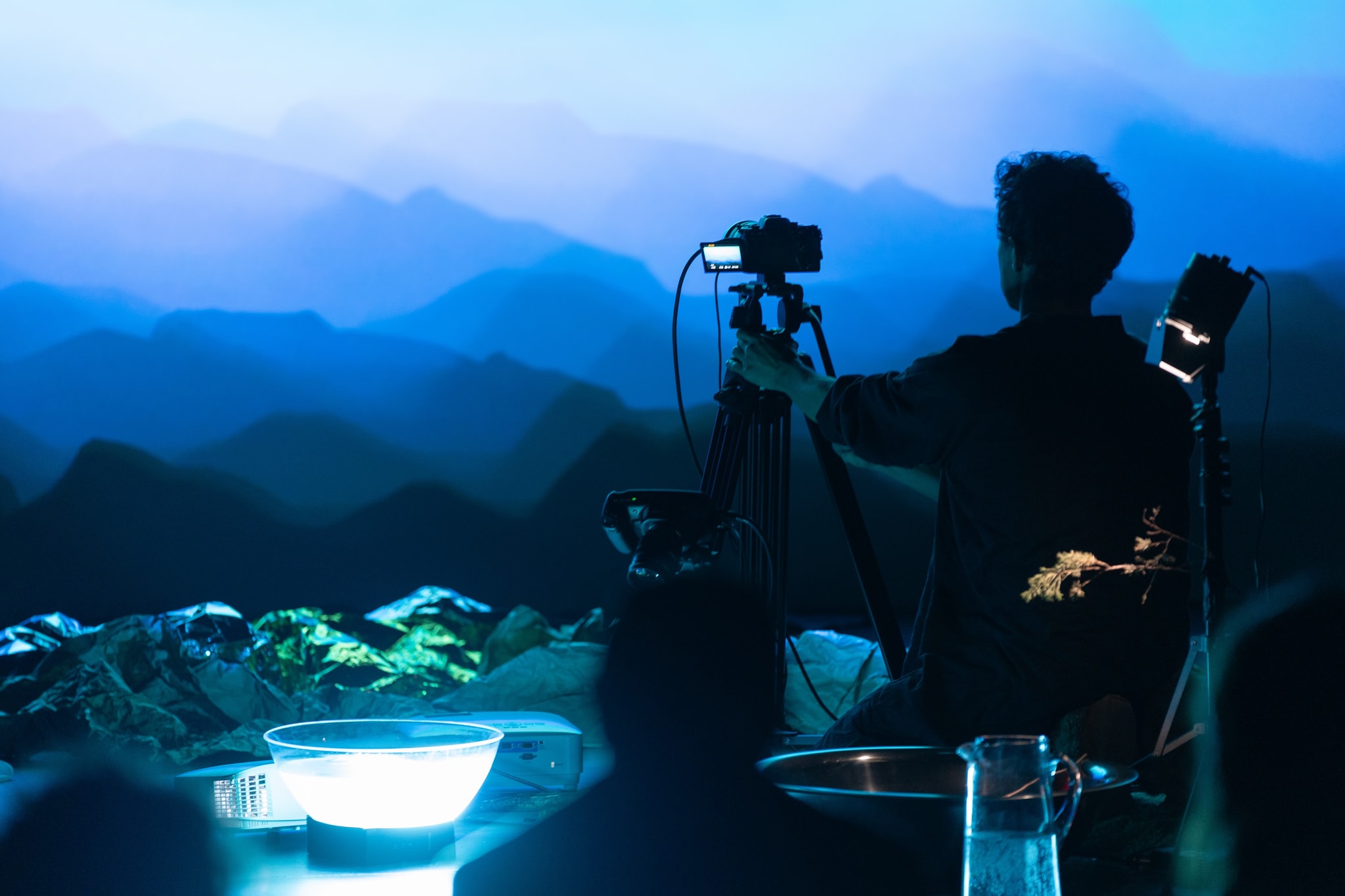 Artist Tyson Houseman is seen from behind in partial shadow as he adjusts a camcorder. Projected in front of him is a blue, mountainous landscape and around him on the floor are small objects, including a glass bowl illuminated by a bright white light. 