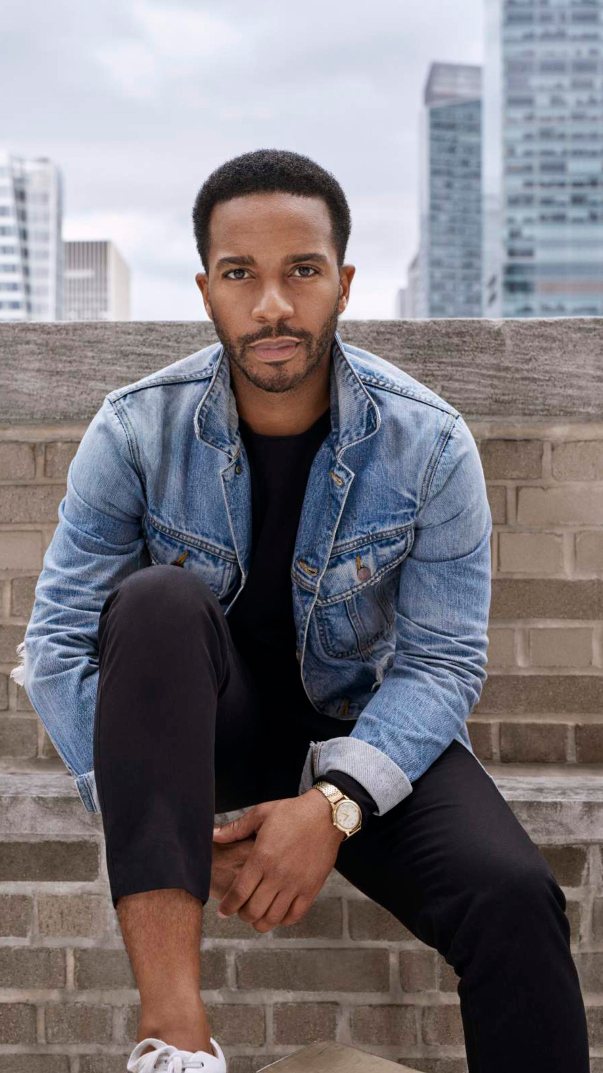 A portrait of actor André Holland. André, who is a Black man with short dark hair and a mustache that connects to his beard, looks intently at us while sitting outside with skyscrapers visible behind him. He wears a jean jacket and leans forward as he has one knee raised toward his chest, clasping his hands under the raised leg. 