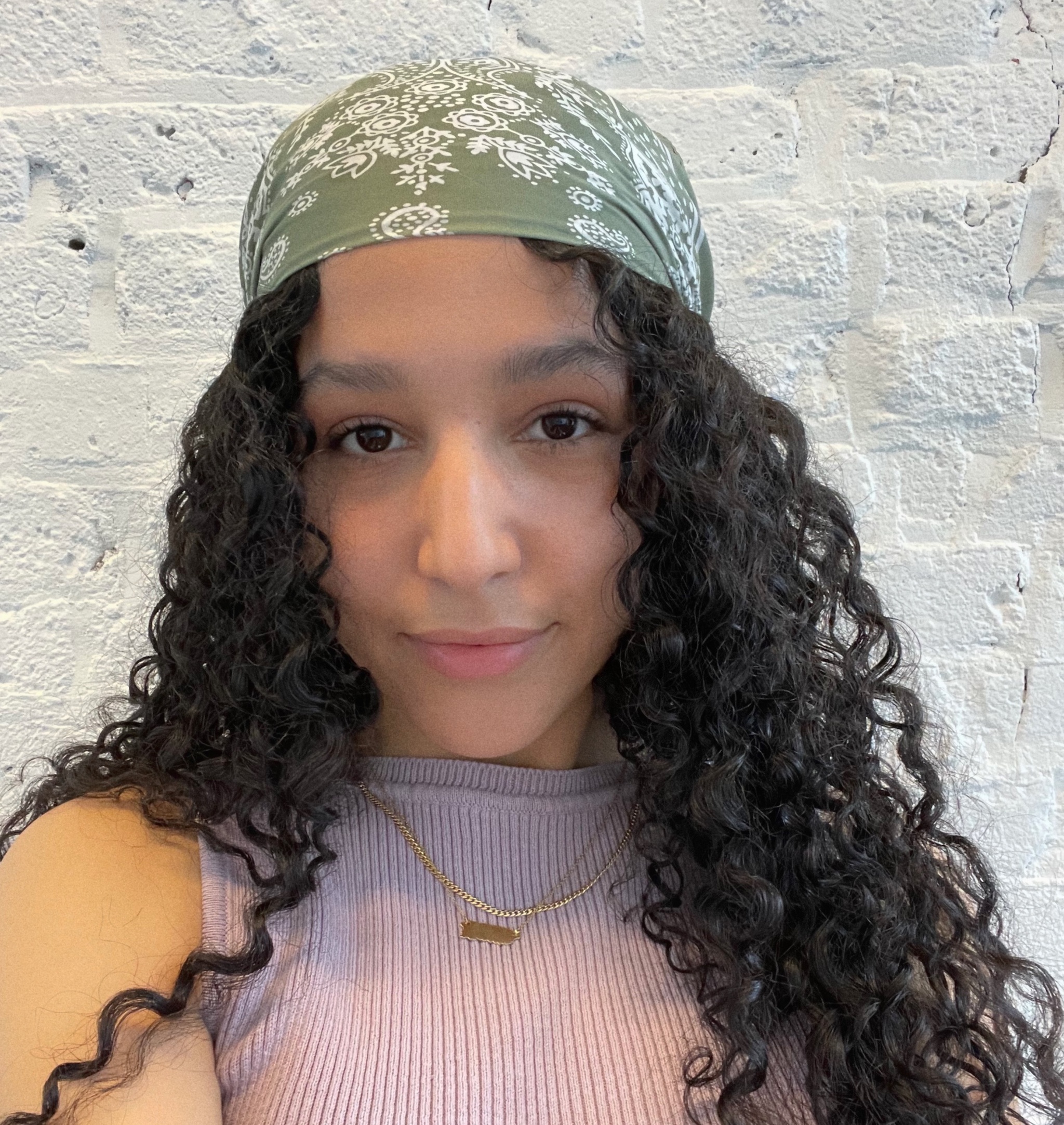 A headshot portrait of artist Mel Corchado. Mel is a Boricua woman whose wavy brown hair flows out of a green scarf she wears on her head. She looks directly at us, with lips curled in a smile, and wears a pale purple sleeveless shirt. 