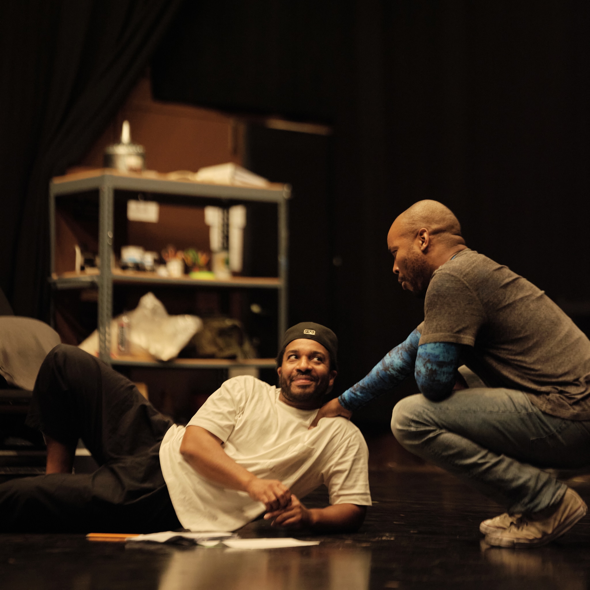 Actor André Holland, a Black man wearing a white t-shirt and baseball cap, reclines on the floor of a black box rehearsal theater. He looks up at Alani iLongwe, another Black actor, who squats beside him, reaching out to place a hand on André's shoulder.