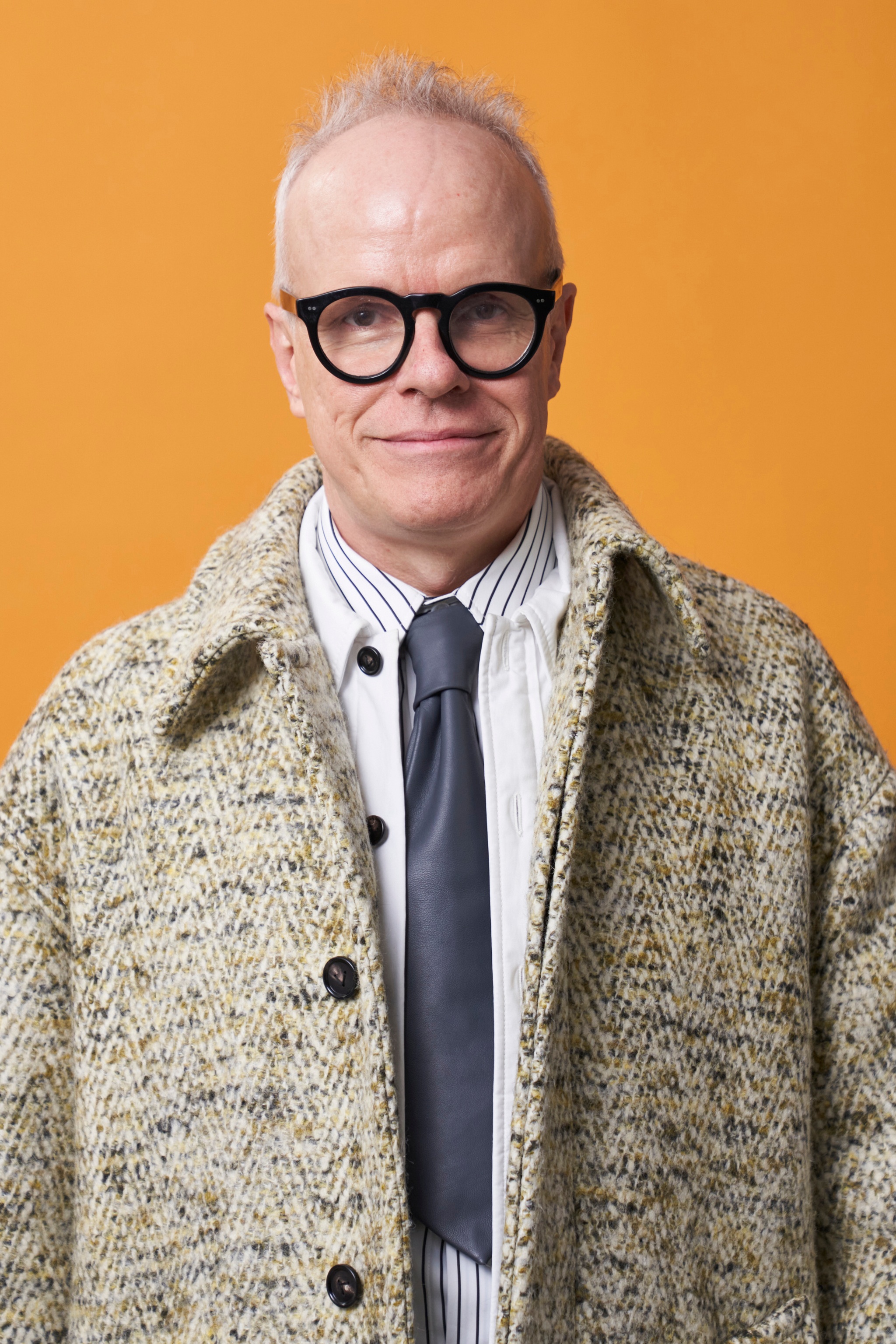 A portrait of curator Hans Ulrich Obrist, a white man with short gray hair who poses against a yellow-orange backdrop. He looks directly at us through thick-framed glasses. He wears a textured wool coat over a shirt and tie. 