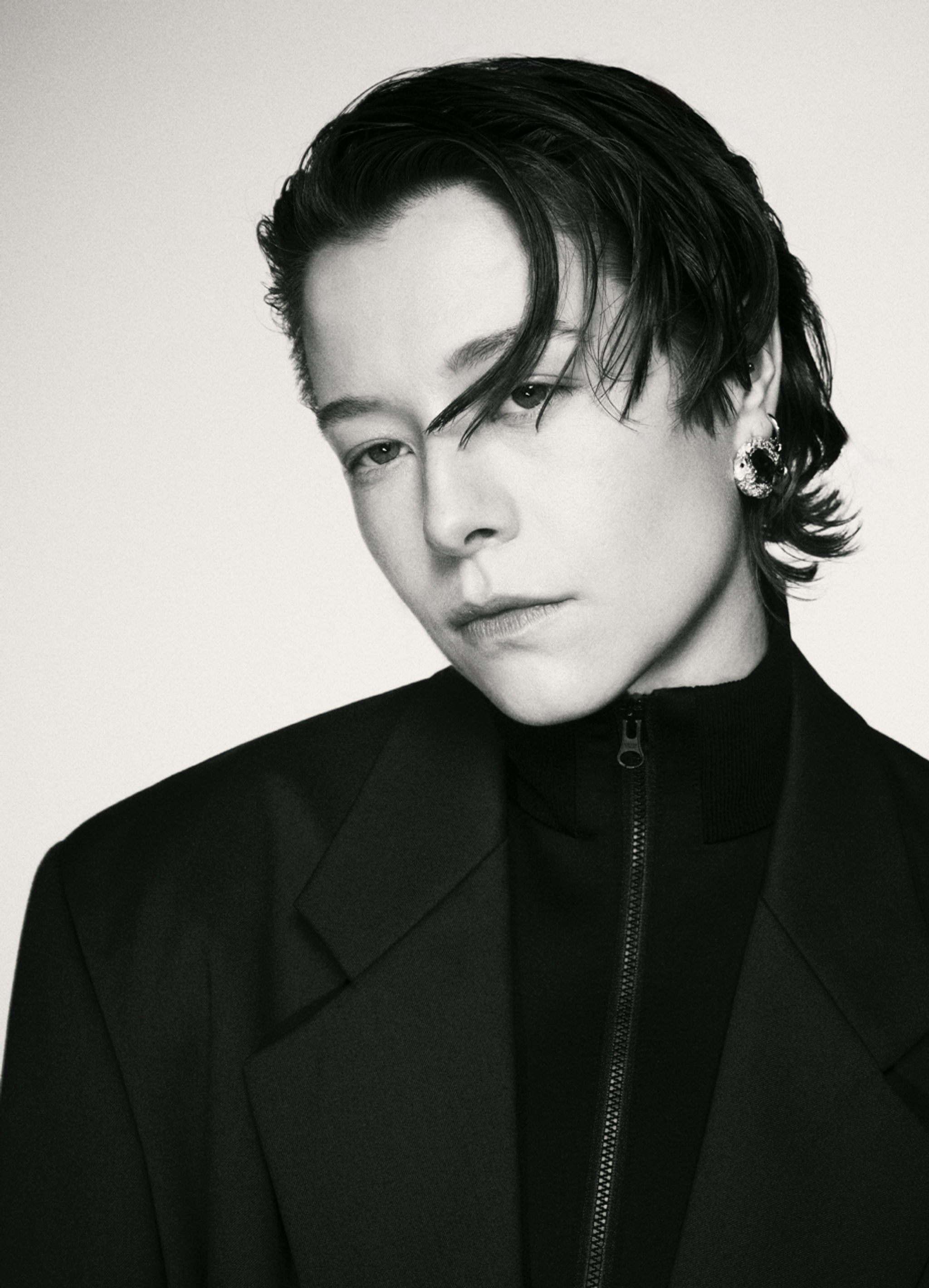A photo portrait of nonbinary actor Emma D'Arcy, in black and white. Emma is a white person with short brown hair styled to swoop down over their left eye. They face us but turn their head slightly at a diagonal away from us. They have a serious expression on their face and wear a blazer over a high-necked dark sweater.