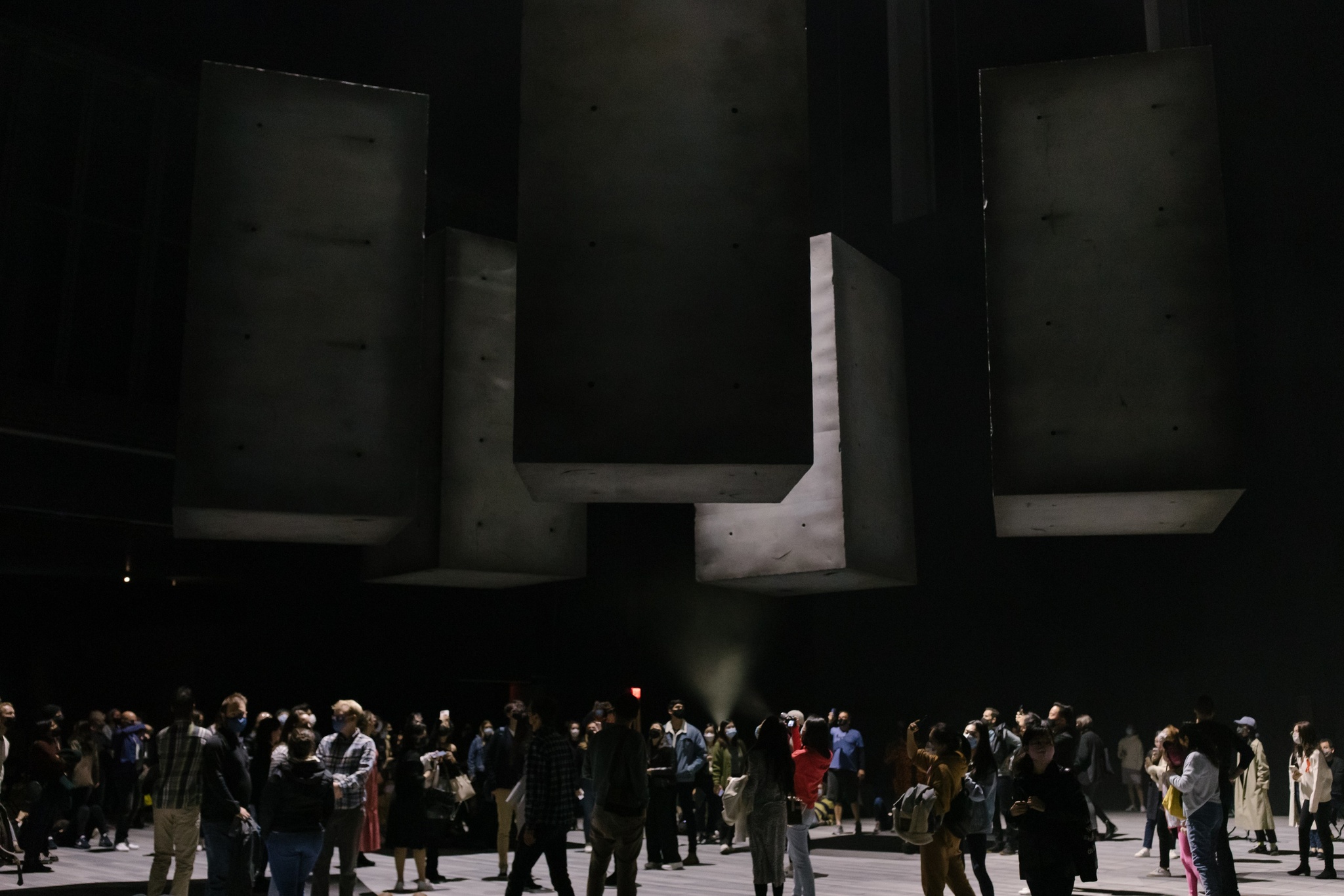 Five concrete blocks float weightlessly in a cavernous, dramatically lit performance space as crowds gather beneath them