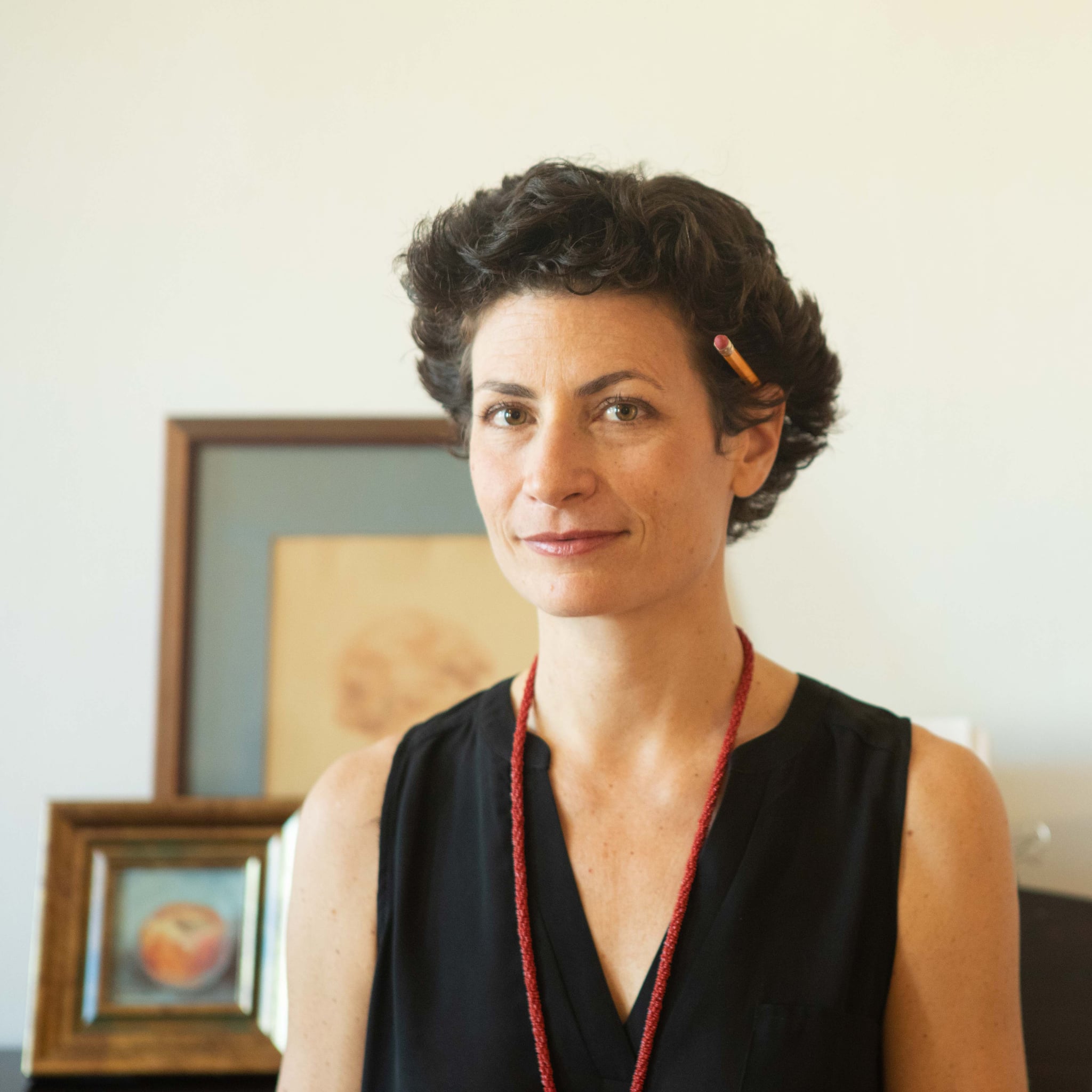 A woman with short hair stands with picture frames propped up on furniture in the background. She wears a deep v-neck top, a long red necklace, and has tucked a pencil behind her ear. 