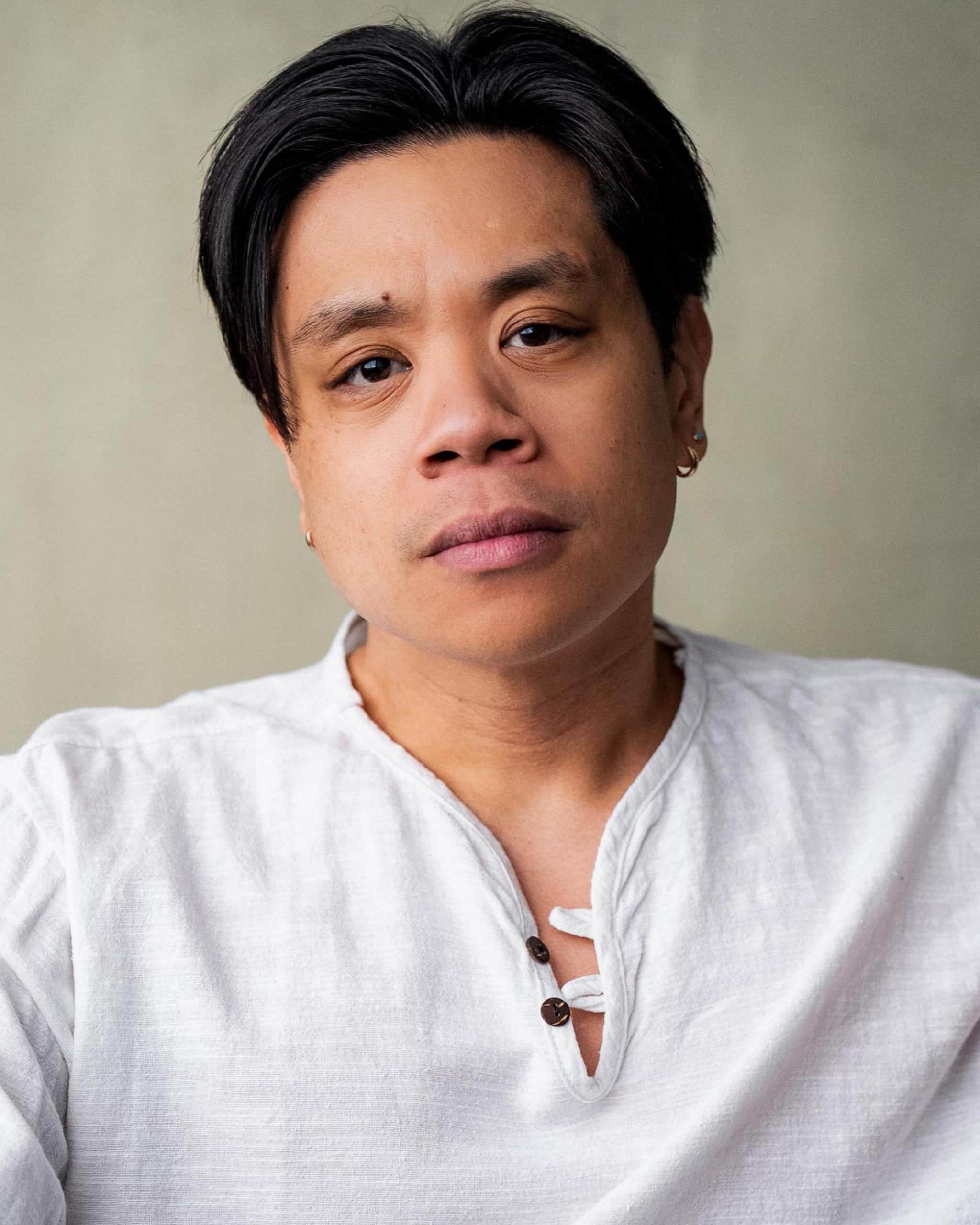 A portrait of Carlo Siriban, who poses against an eggshell background. He is seen close up, from chest up, and looks directly at us with a calm but serious expression. His dark hair is parted in the center so that it falls away from his forehead. 