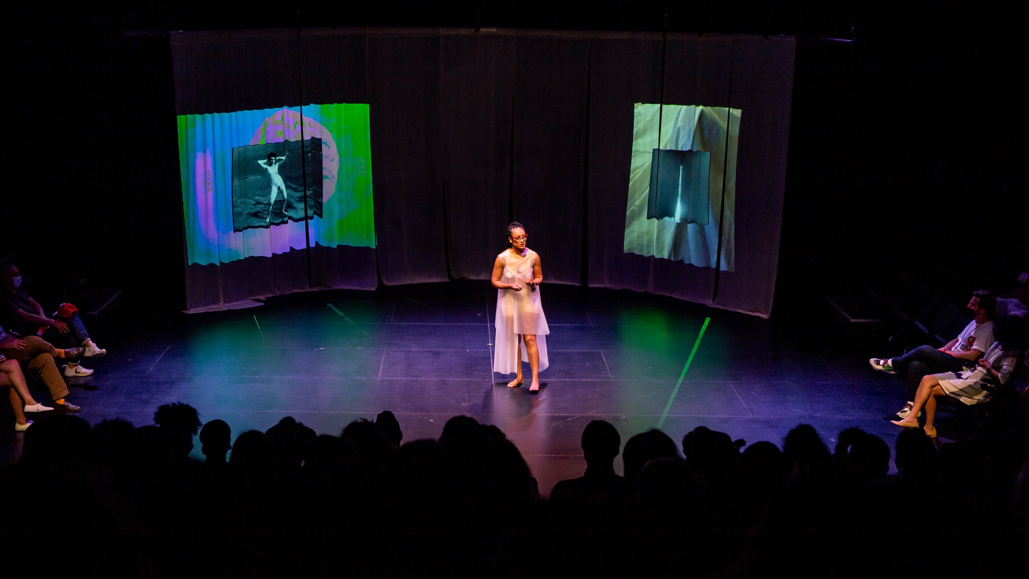 The artist Eleanor Kipping performs on stage in a darkened theater in front of an audience. She wears a sheer pink dress and red framed glasses. She is speaking to the audience, flanked by two screens with video projections.