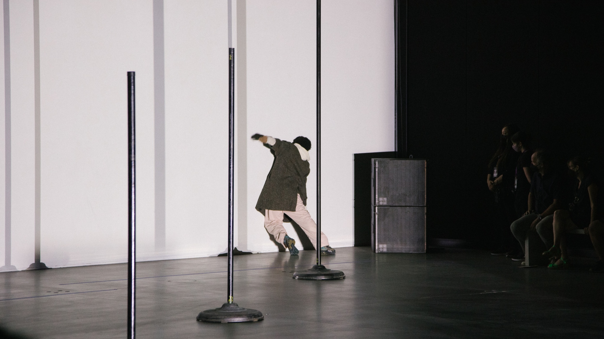 The artist Niall Jones leans the weight of his body against a white wall during a performance so that he is supported by the force of his body. He is facing the wall so his face is not visible.