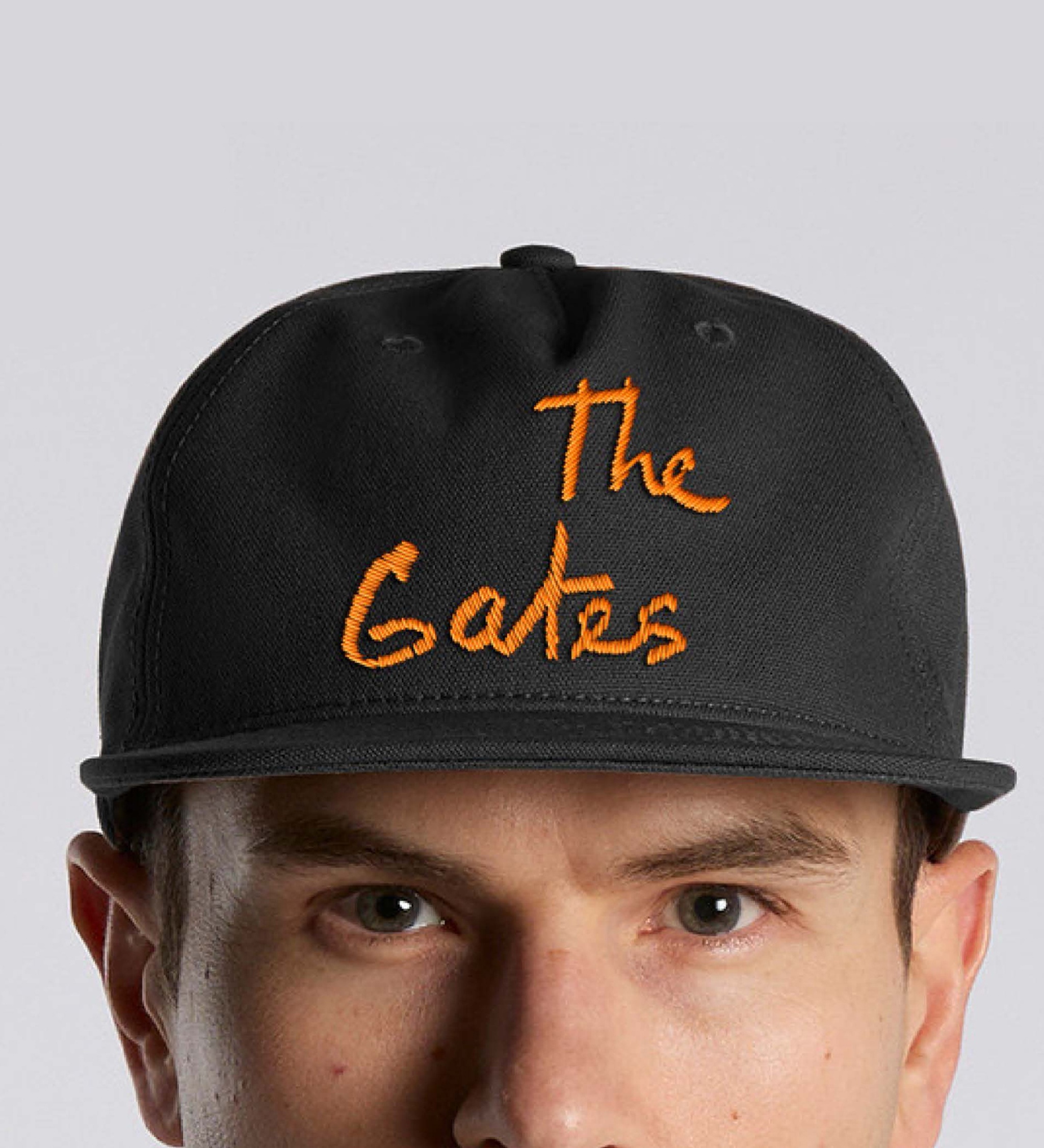 A black canvas cap with the artwork title The Gates in saffron embroidery above the bill