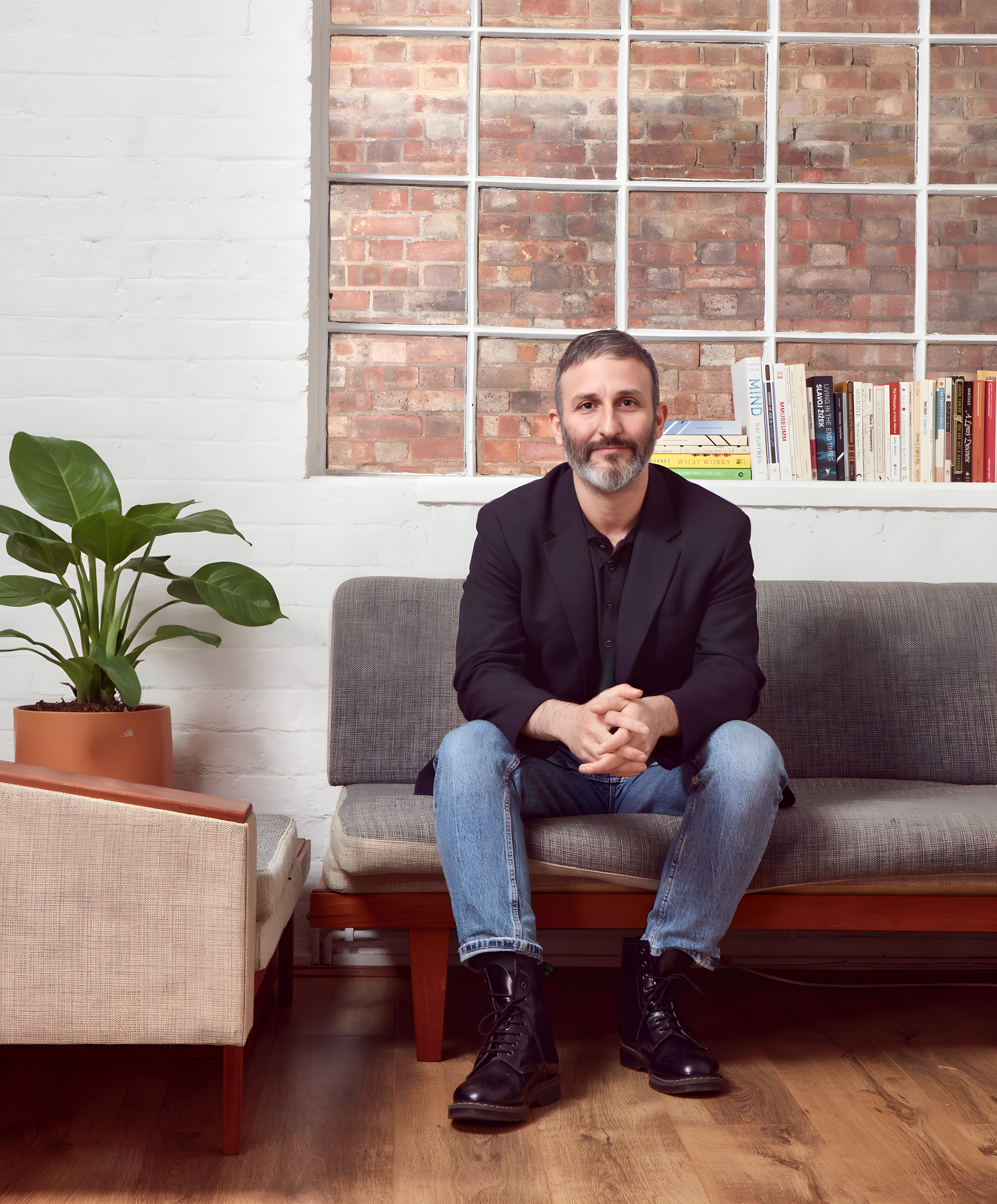 A portrait of artist Igal Nassima. He sits on a gray sofa with hands clasped between his knees. He wears light blue jeans and a darker blue shirt. She has a grayish beard and looks directly at us. Behind him is a brick wall visible through a large window. 