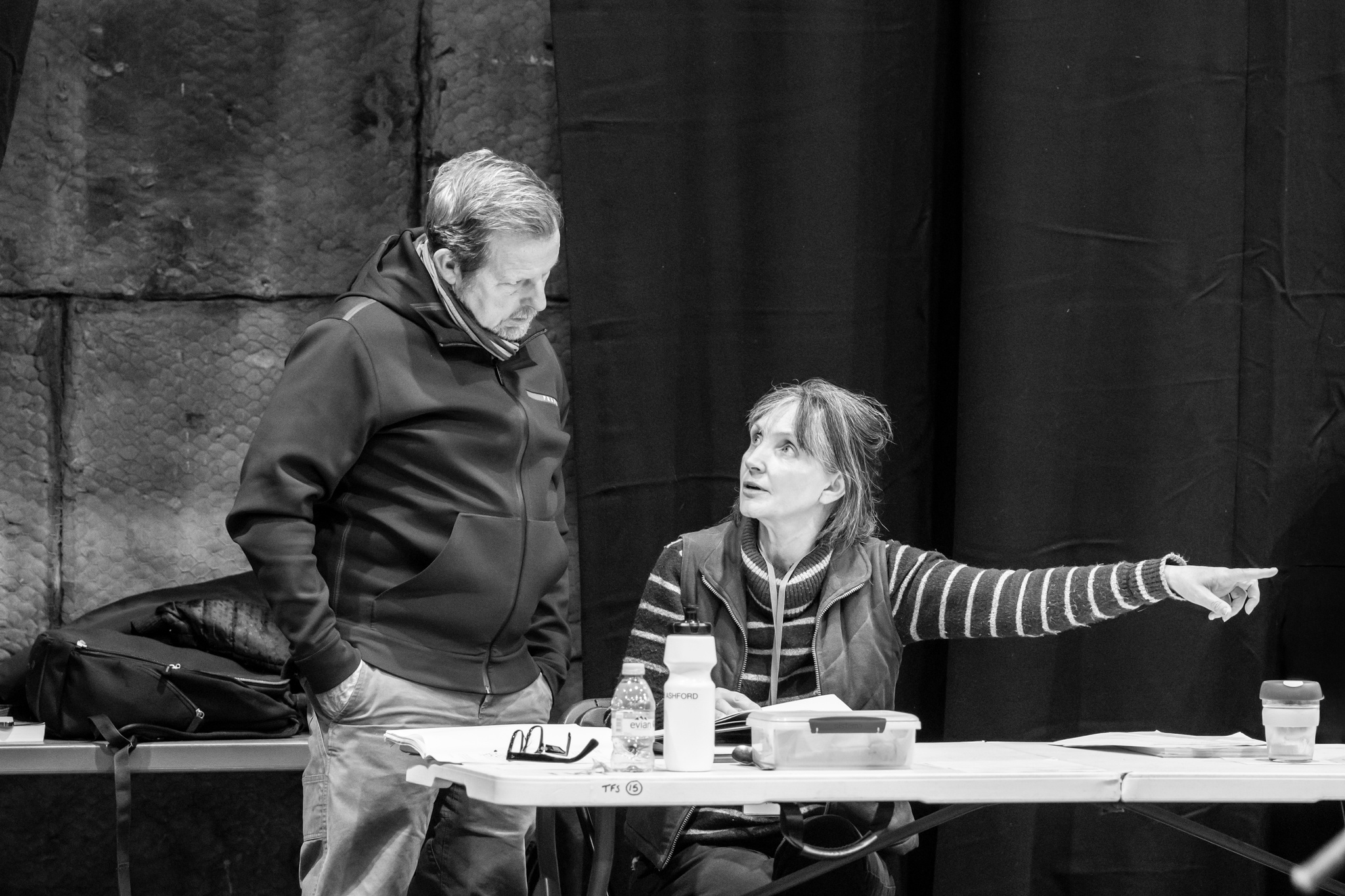 Co-directors Rob Ashford and Lucy Skilbeck confer during a rehearsal. Lucy sits at a table pointing to her side, while Rob looks down at the papers on the table in front of her.