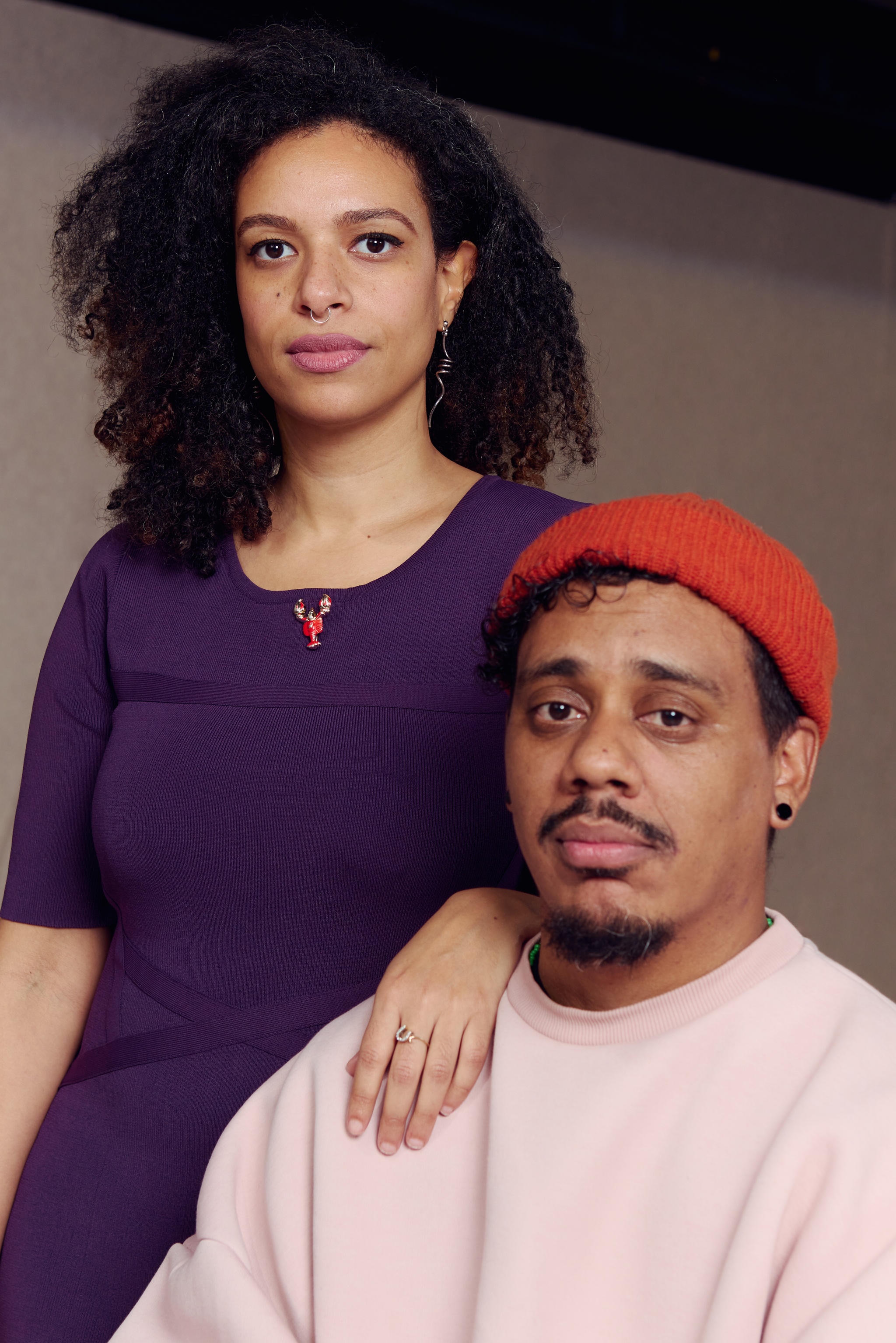 Artist duo Yelaine Rodriguez and Luis Vasquez La Roche pose together. Yelaine in a purple dress stands behind Luis, who is seated, with one hand on Luis's shoulder. Luis wears an orange beanie and a pale peach sweatshirt. They both look directly at us. 