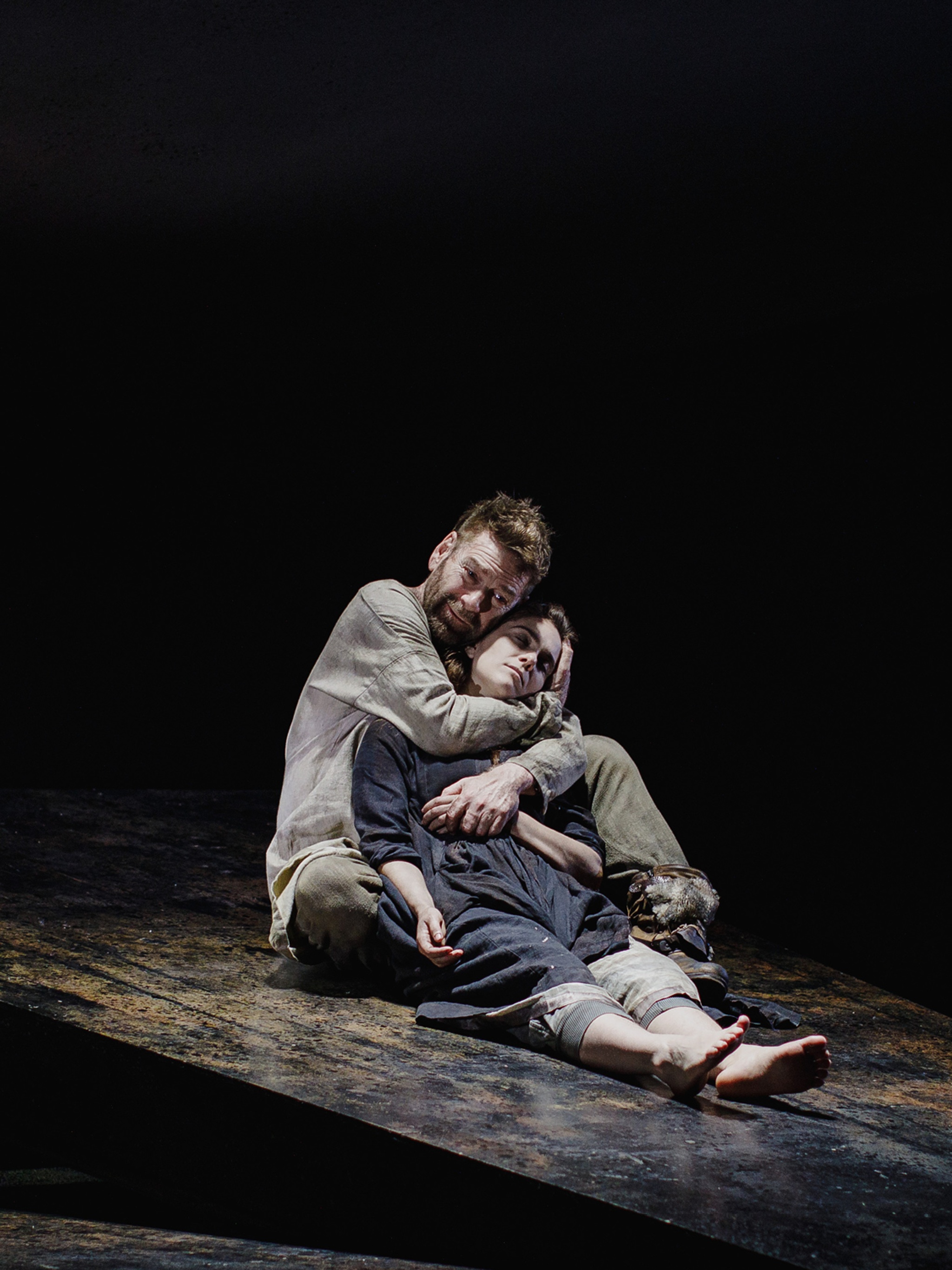 A production photo of actors Kenneth Branagh and Jessica Revell on stage during King Lear. Both are on the floor of the stage as Branagh, playing Lear, cradles Revell, who is playing Cordelia, in his arms. Branagh's face is griefstricken while Revell looks blankly after her character has died. 