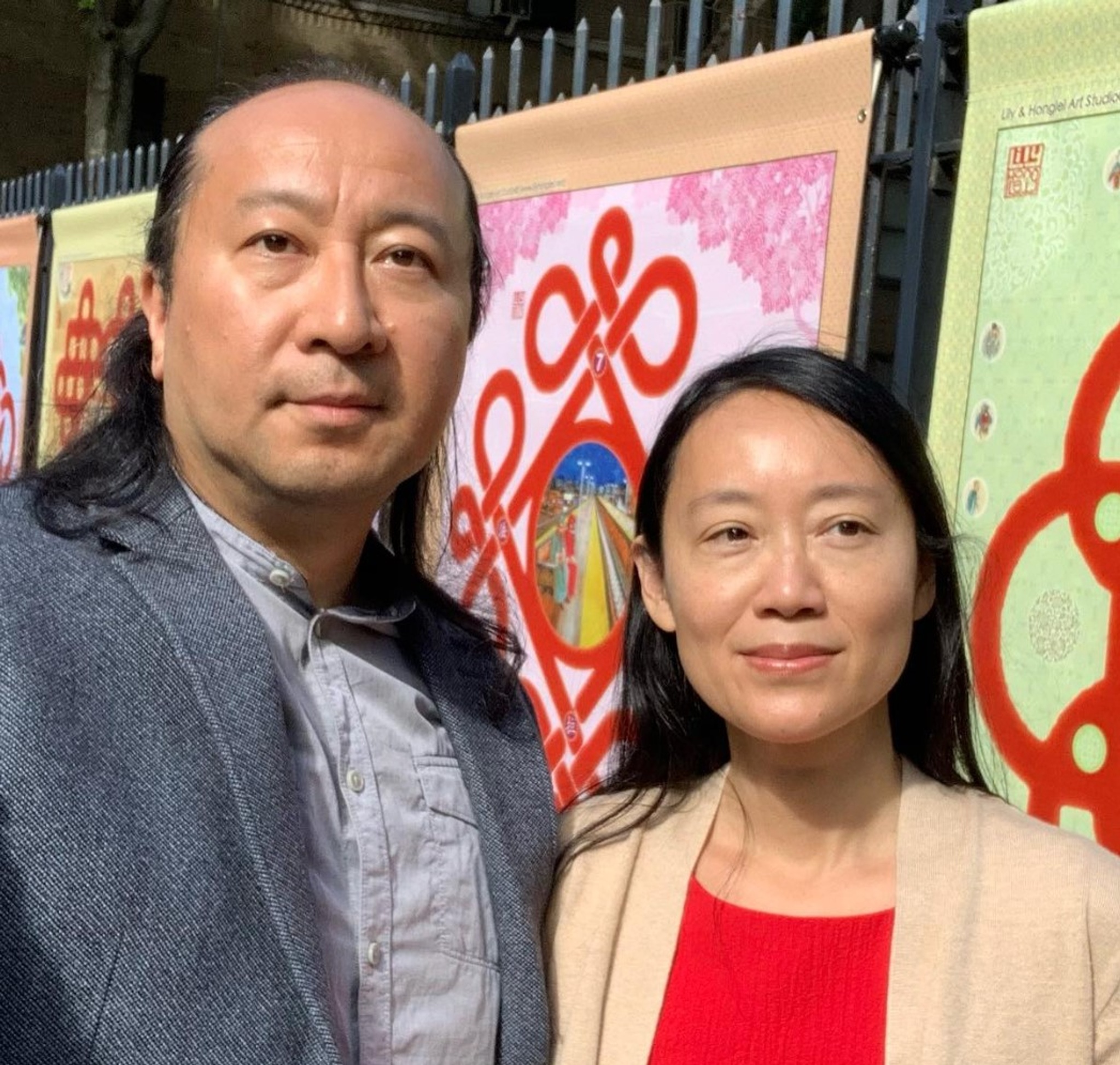 A headshot of the Chinese American artist duo Lily Honglei. They stand shoulder to shoulder, behind them are colorful banners hanging from a fence. 