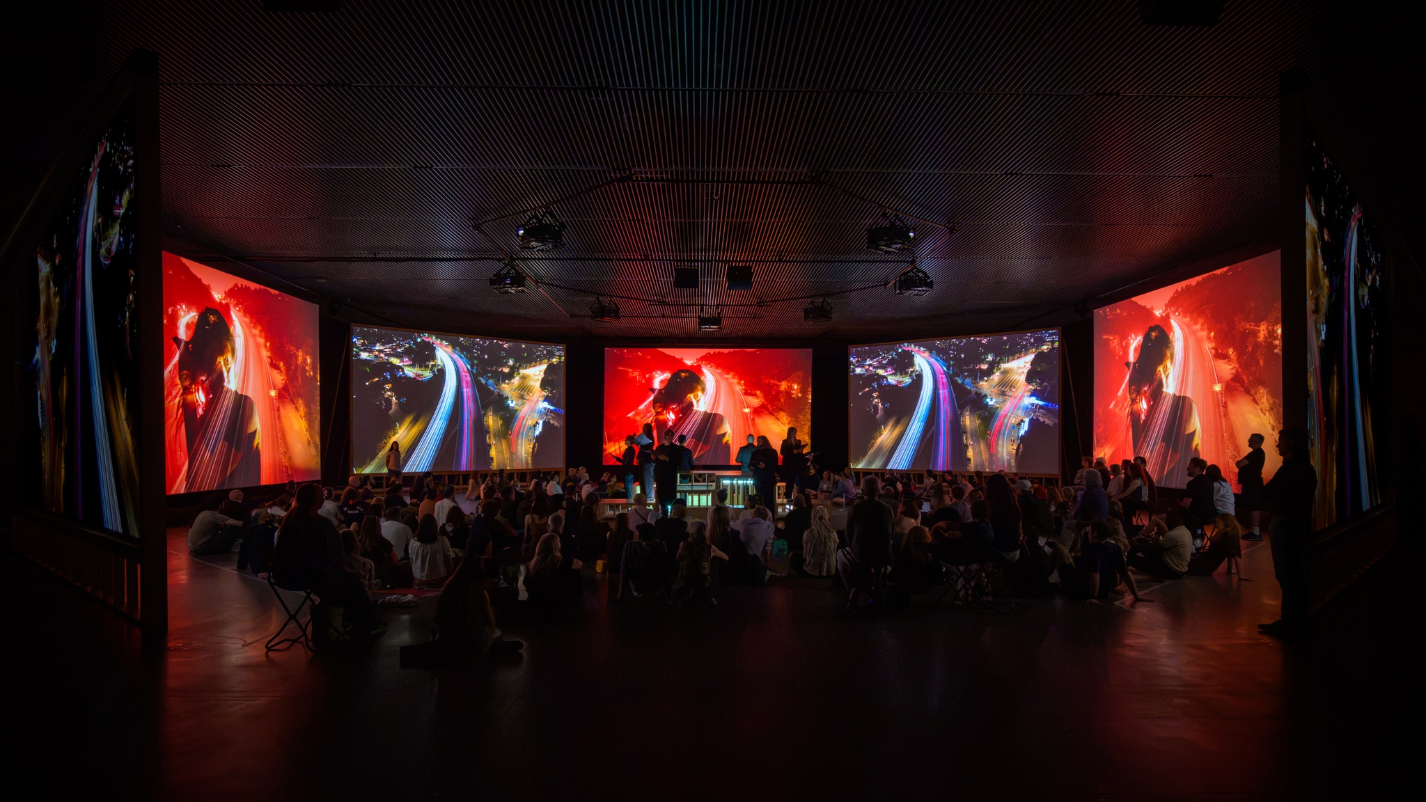 A group of chorus members dressed in black sit on a low platform in a gallery surrounded by film screens and audience seated on the floor around them.