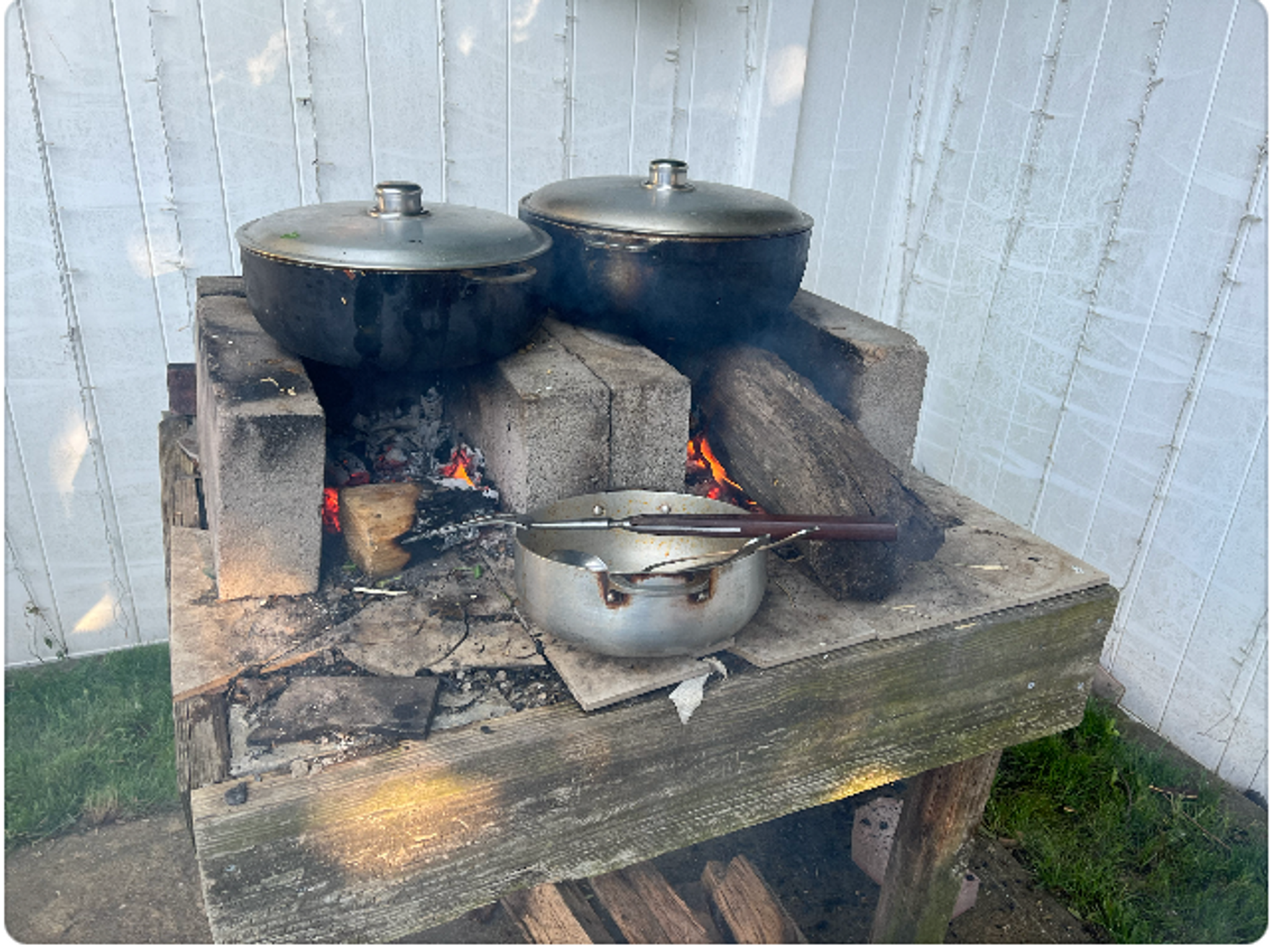 A makeshift backyard barbecue with two heavy iron covered pots sitting on a fire that is glowing orange beneath them.