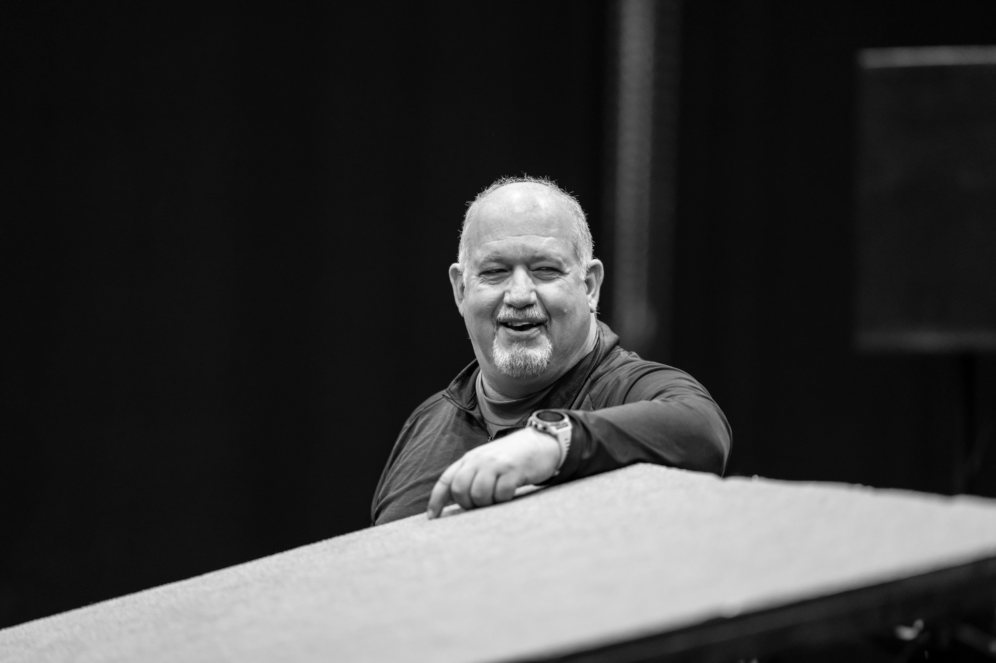 Fight director Bret Yount pauses during rehearsal. He is smiling and looking off to his side, with one hand casually resting on a platform in front of him. 