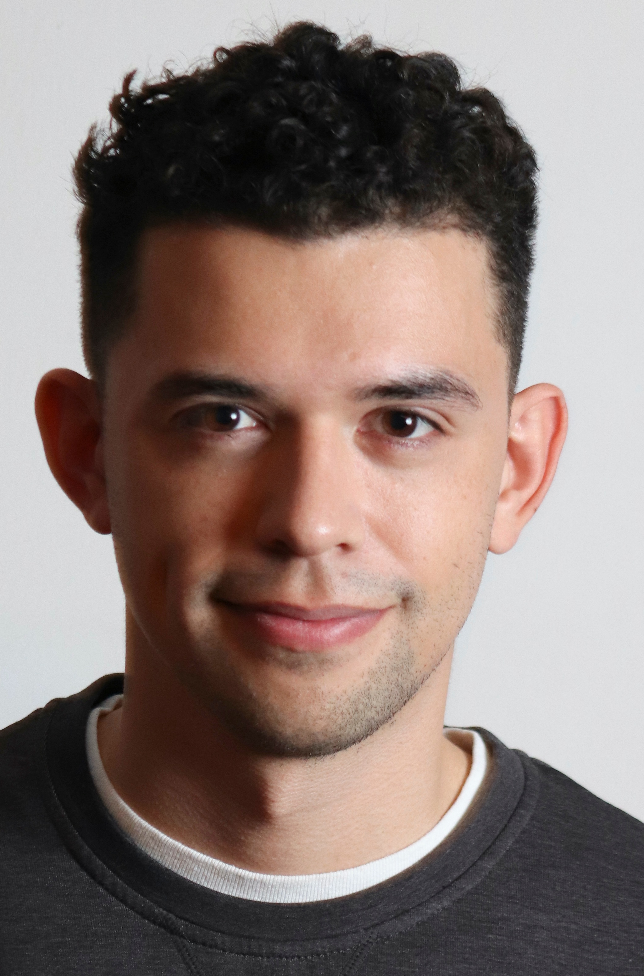 A portrait of dramaturg Jeffrey Gan. He has dark brown hair that curls on top with the sides of his head buzzed closer to the scalp. He grins broadly without opening his mouth. 