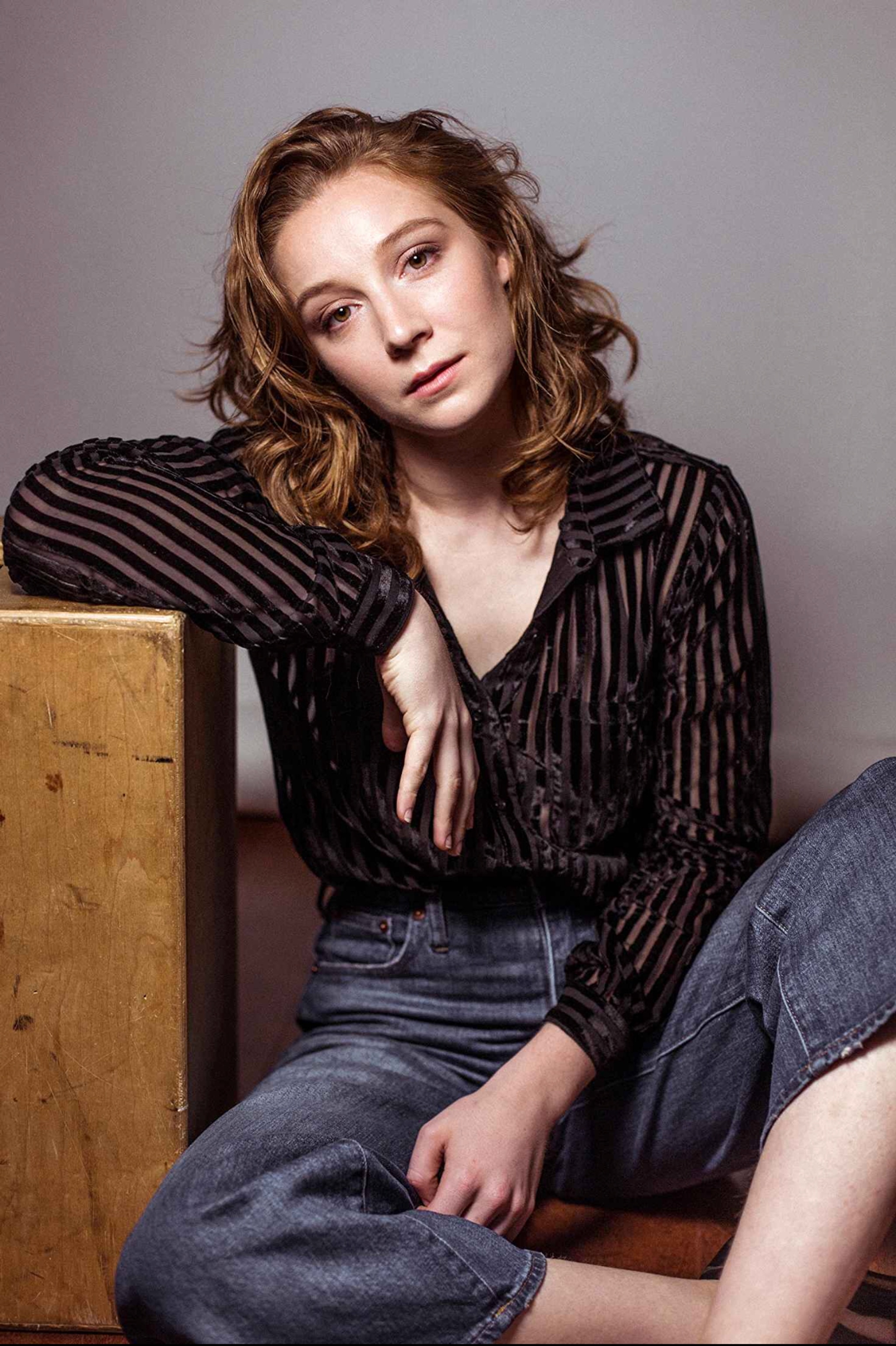 A studio portrait of Kayli Carter, a white woman with reddish brown hair who sits in a relaxed pose against a wooden box. She leans on the box with one arm propped on it and her legs drawn up toward her body. She wears a pair of jeans, a striped sheer blouse and leans her head to the side as she looks at us thoughtfully. 