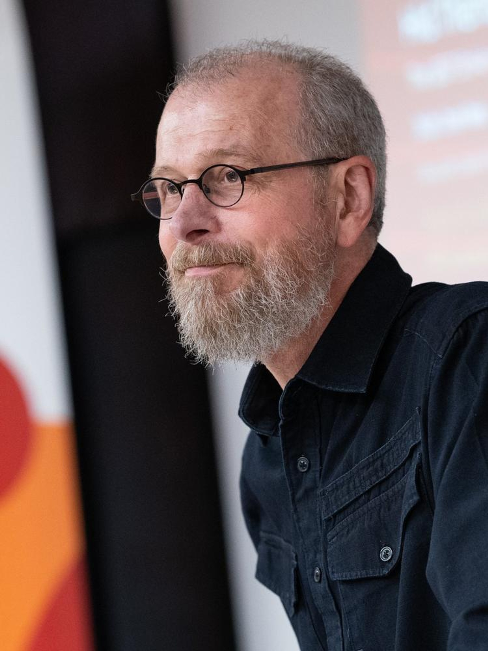 A portrait of João Biehl, a white man with gray hair and beard and mustache. He is seen at a presentation in three quarter view, leaning slightly forward with a screen in the background. He wears glasses and a blue collared shirt. 