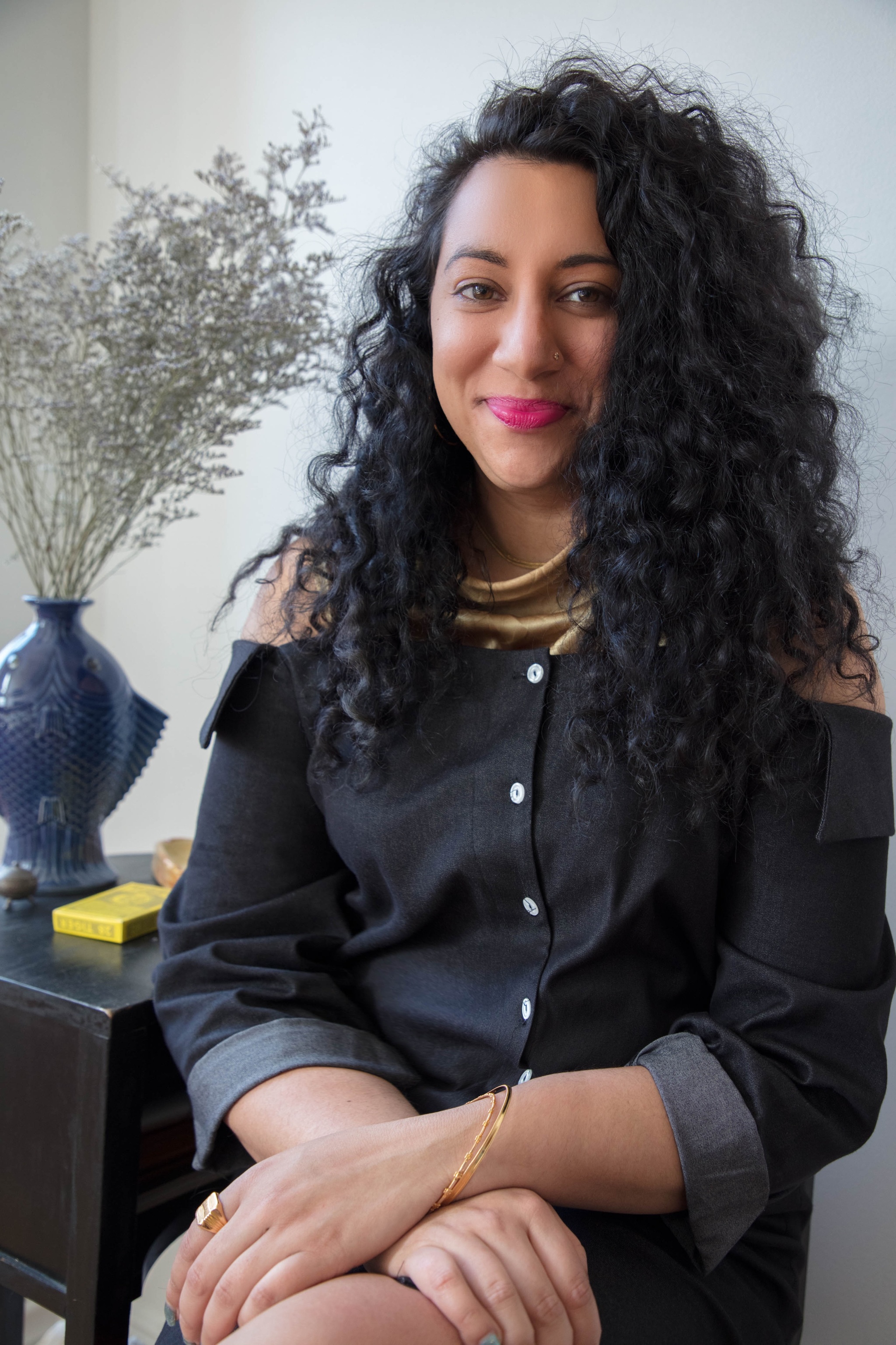 A photo of Diya Vij seated with hands crossed atop her leg. Vij has long, curly hair parted to the left. 