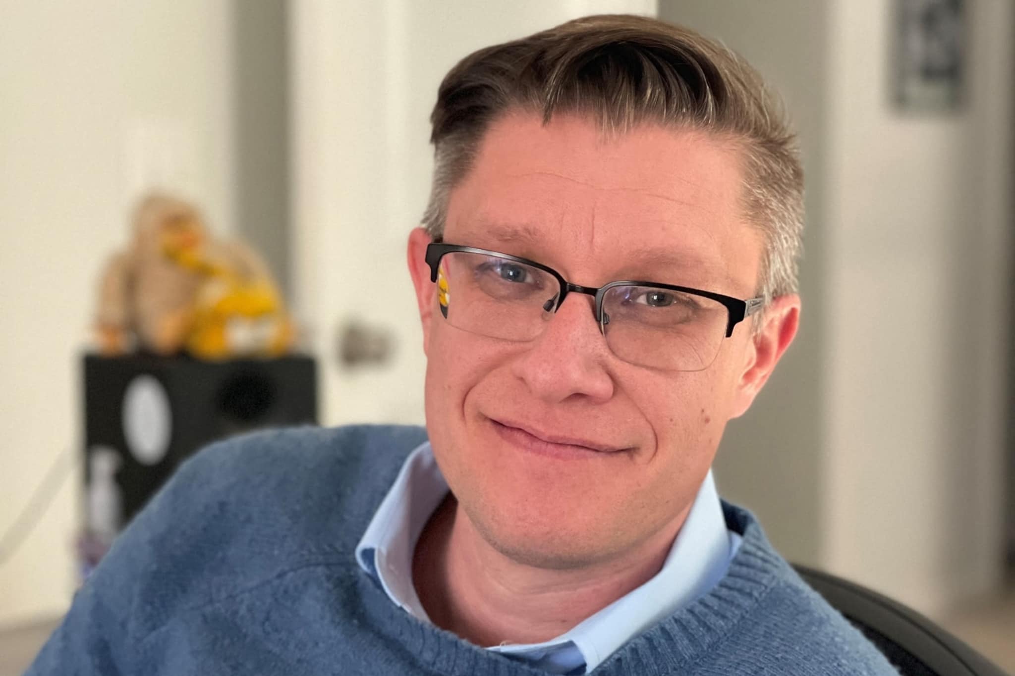A portrait of digital artist Beeple, a white man with salt and pepper brown hair neatly parted to the side. He wears glasses and a blue sweater with a shirt collar. He smiles broadly at us. 
