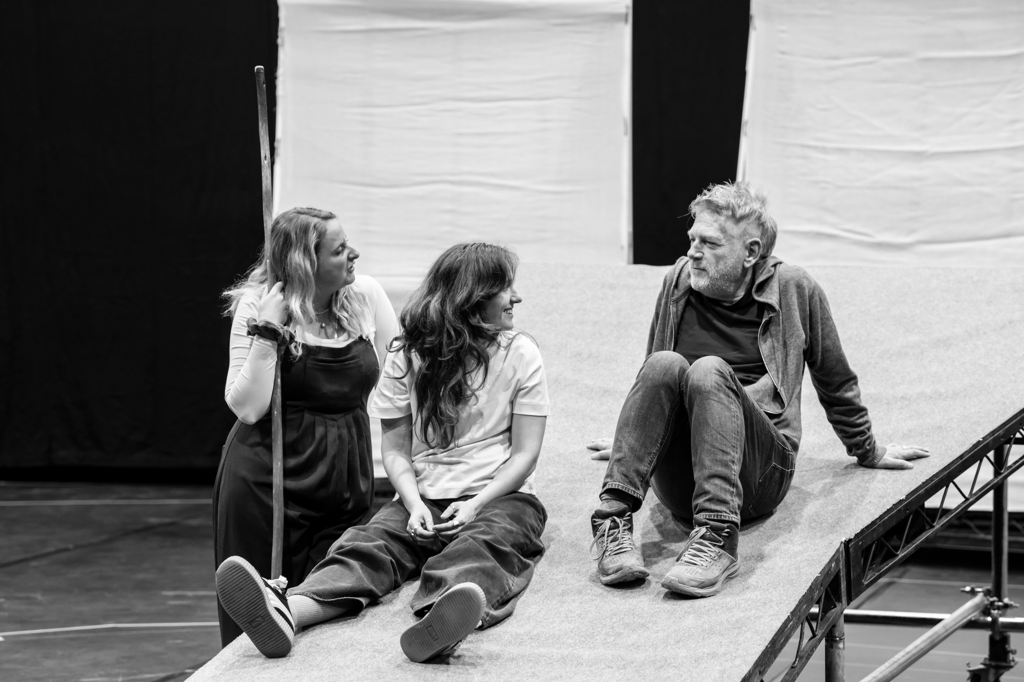 Actors Eleanor de Rohan, Jessica Revell, and Kenneth Branagh relax during rehearsal. Kenneth and Jessica sit on a platform, while Eleanor stands leaning on it. Jessica and Eleanor look toward Kenneth with smiles on their faces.