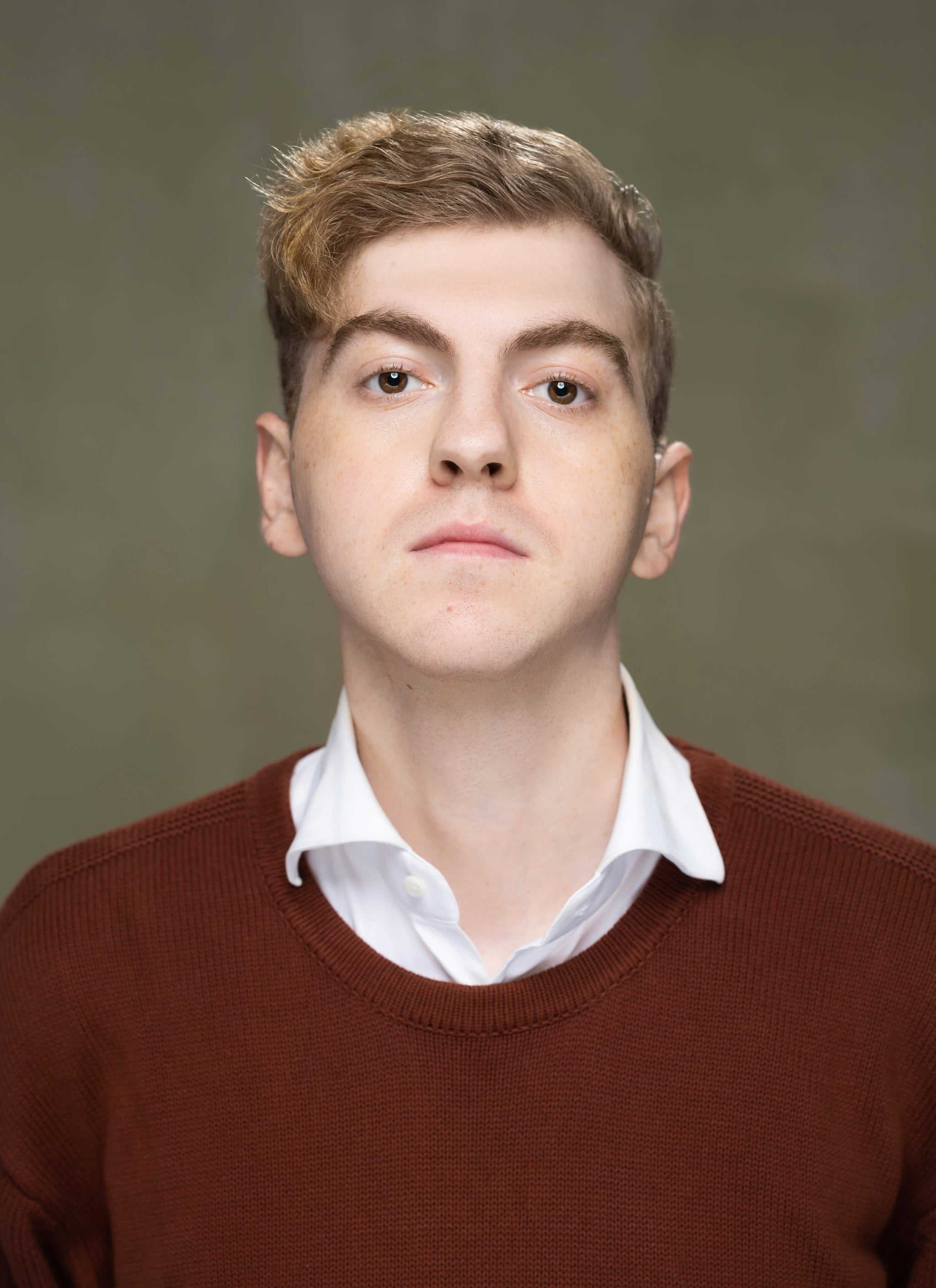 A portrait of Samuel Langshteyn, a white Deaf actor, who is seen from the chest up against a neutral backdrop. He wears a burgundy sweater with a white collared shirt popping out from the neckline. His dirty blond hair is swooped across his forehead and he looks directly at us with eyebrows raised,