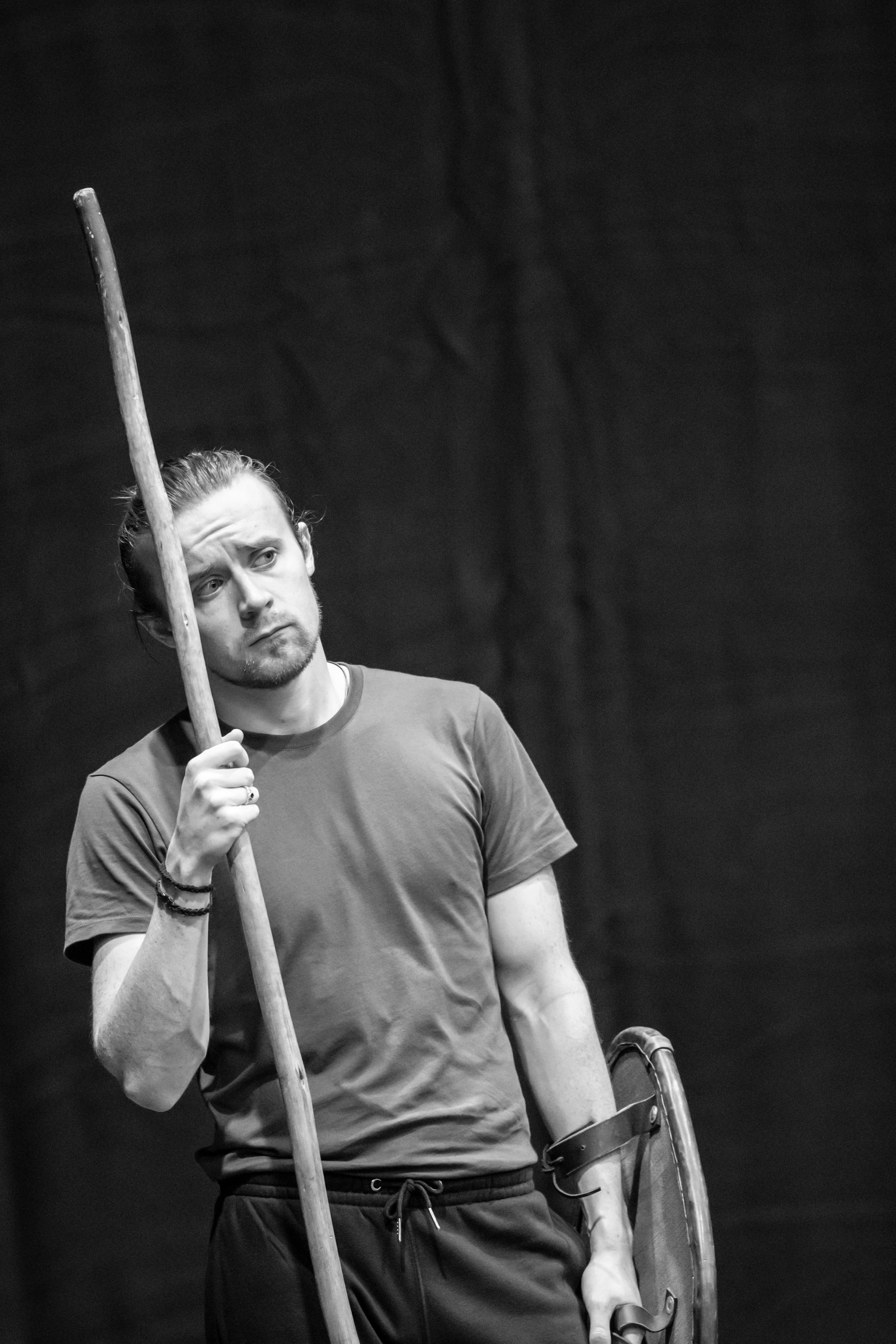 Actor Stefan Brennan-Healy, a white man, stands holding a large staff in rehearsal. He wears a gray tshirt and is looking to the side with a concerned look on his face. 