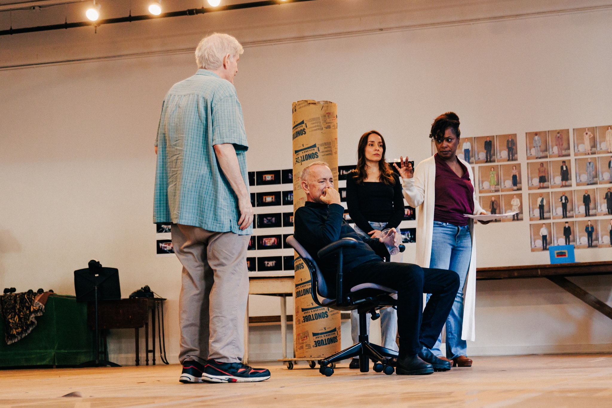 Actors Paul Murphy, Tom Hanks, Jamie Ann Romero, and Michelle Wilson rehearse in a dance studio space. Tom is seated while the others stand around him with their attention on him. Michelle gestures energetically to him as if making a strong point in conversation. 