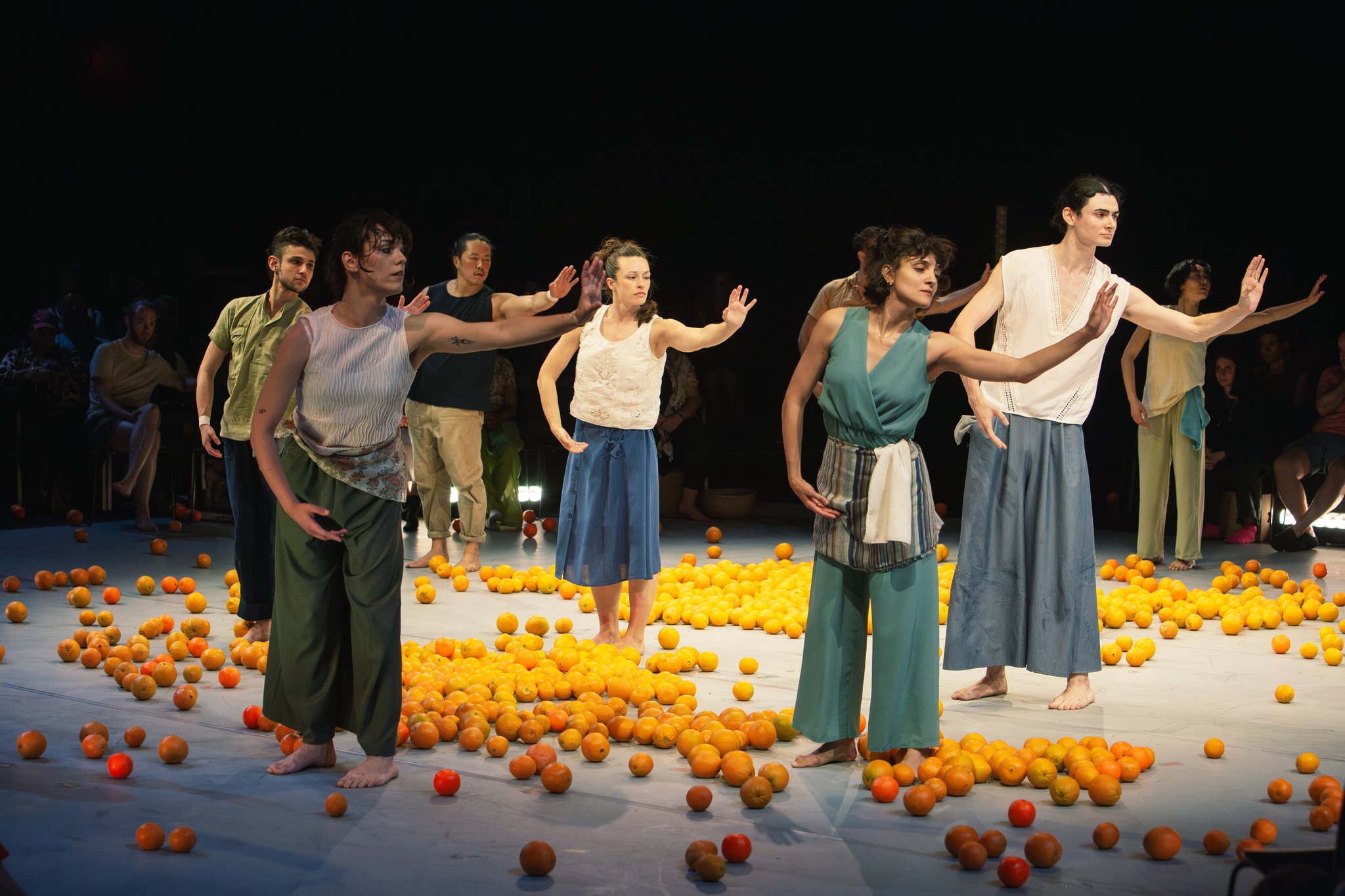 Seven dancers stand in a roughly circular formation in a spotlit stage area. They stand with their left arms gracefully outreached, leaning slightly in that direction. Around them are scattered bright oranges on the stage. 