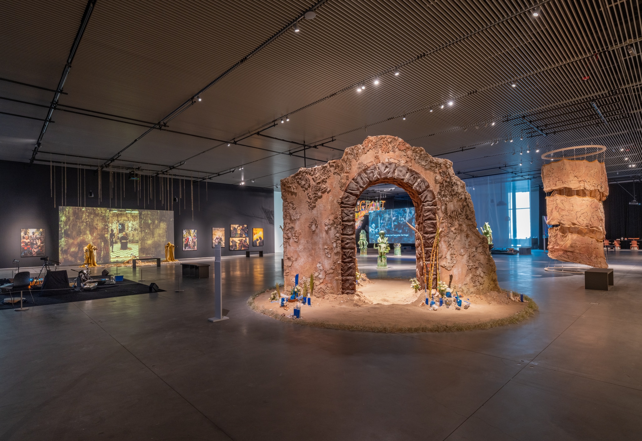 An installation view of a gallery exhibition. The gallery is a cavernous space without walls and cast in dim light. At the center stands mysterious ruined archway stands 12 feet tall in a gallery space. The coarse surface of the archway, built with drywall, has a rough, distressed texture as it is, in fact, covered with a mix of ground coffee, tobacco, and sugar, disguising the drywall as if this archway were built from ancient stone.