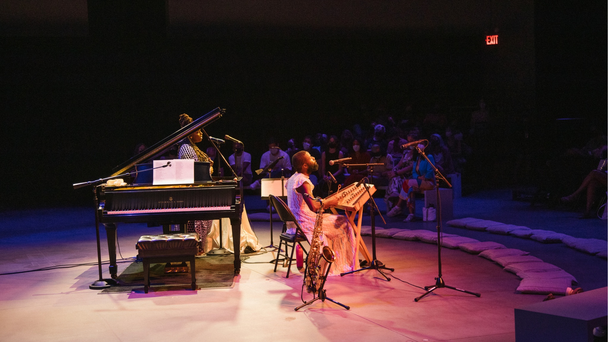 The artist Jerome Ellis sits in front of an audience arranged in a semicircle in front of him in a darkened theater space. Jerome is a Black man who wears a long dress and has a beard. Beside him is a baby grand piano and a Black woman who is a singer.