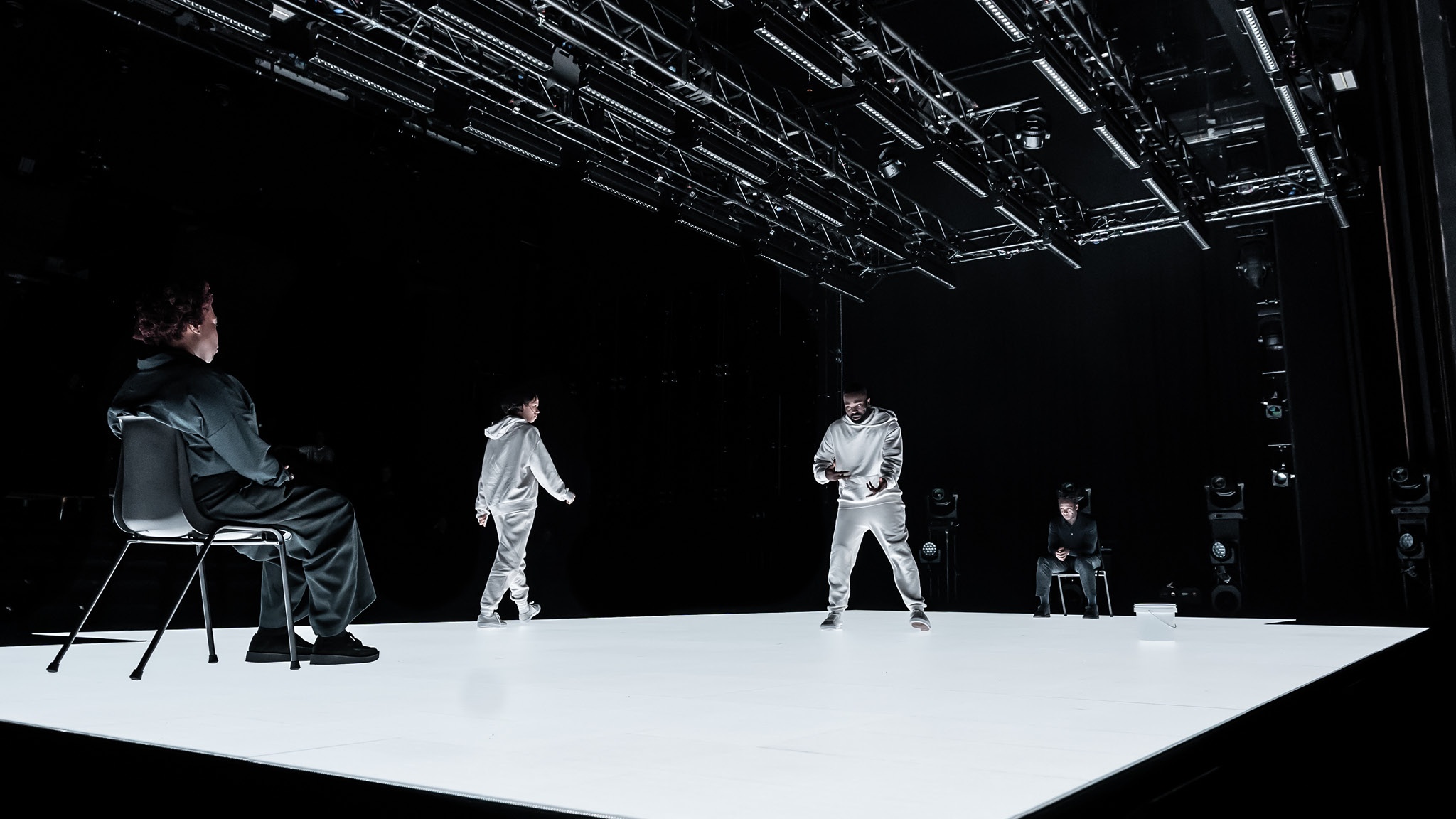 The actors Michele Austin, Taylor Russell, Paapa Essiedu, and Kobna Holdbrook-Smith on stage in The Effect. The stage is a platform lit like a light box, giving off a white glowing light. Michele and Kobna are seated at either end, while Taylor and Paapa pace around each other wearing white sweatsuits in the center of the stage.